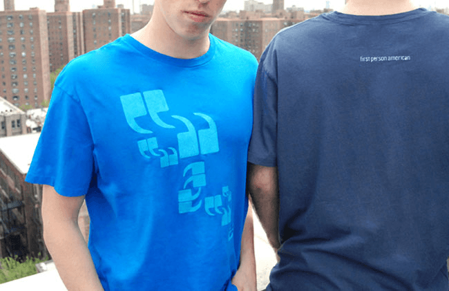 Two people are standing close together. The person on the left wears a bright blue t-shirt with a light blue design that includes quotation marks and stylized symbols. The person on the right wears a dark blue t-shirt with the words “first person american” printed on the back. The background shows a cityscape with tall buildings.