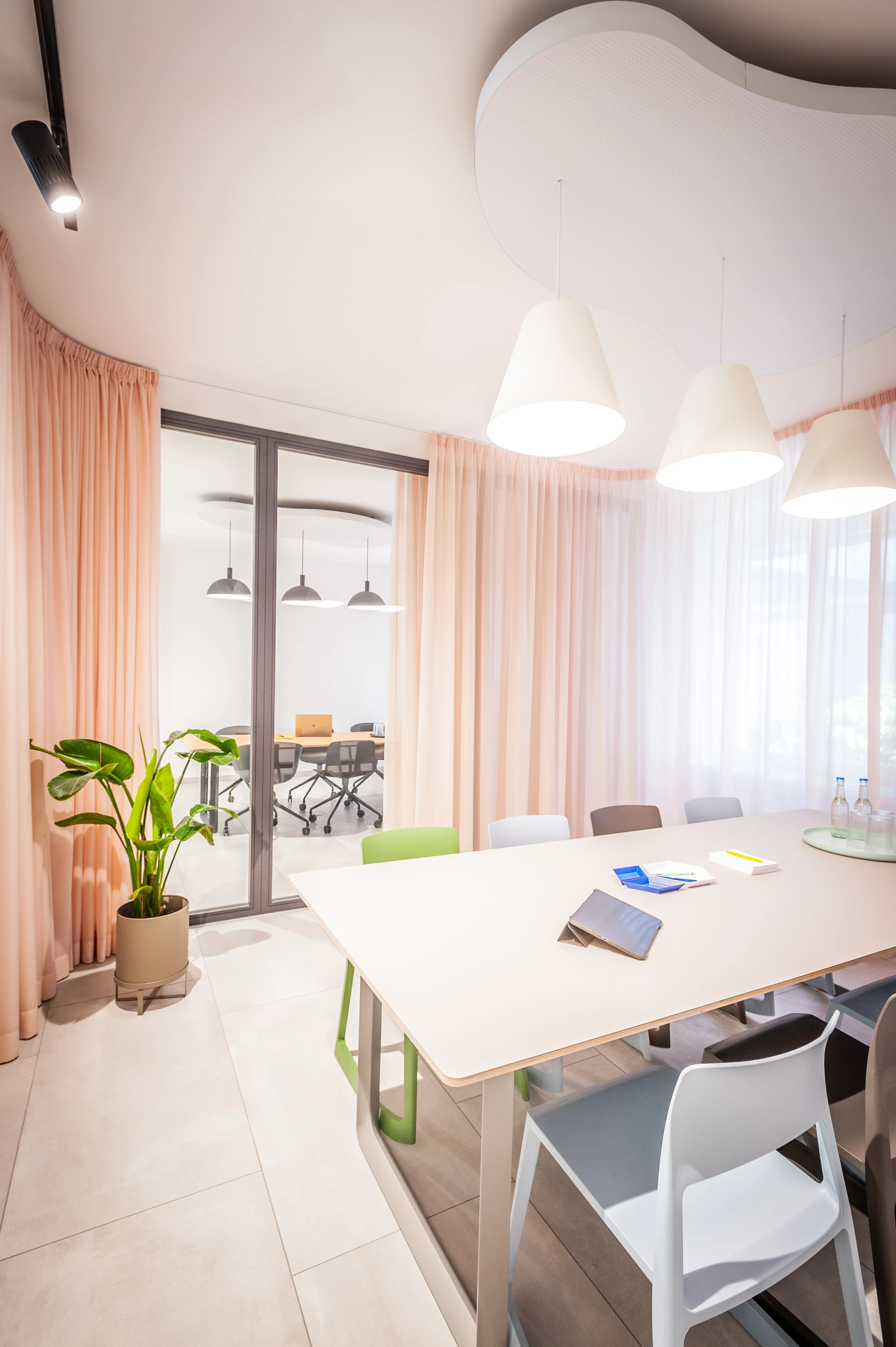 Meeting room at EHEALTH-TEC with a curved white ceiling canopy, white cone pendant lights, floor-length blush-pink curtains, a white table with green and white chairs and a potted plant.