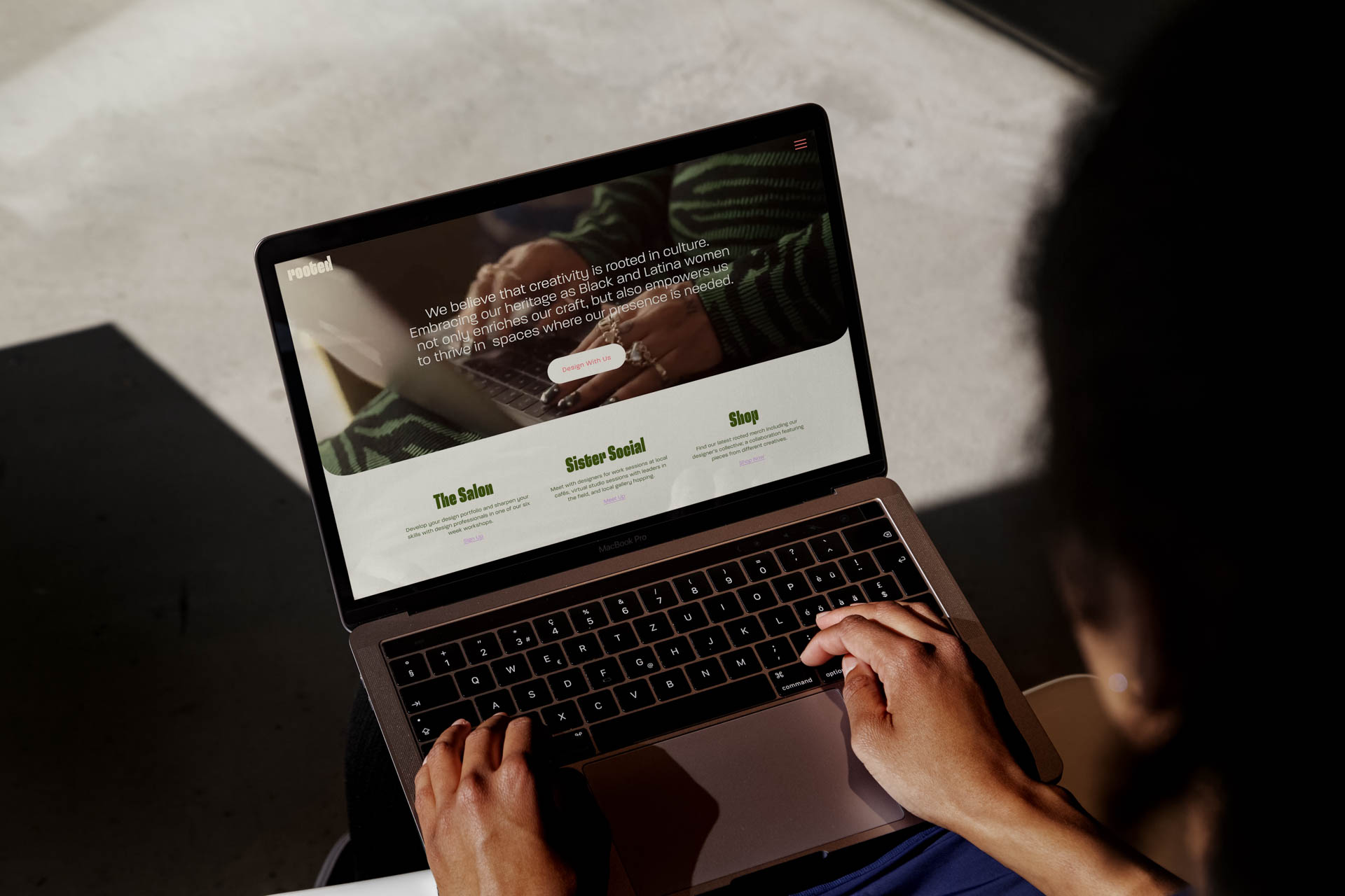 A person is using a silver laptop computer. The screen displays a webpage with the title “Sister Social” and three green buttons labeled “Tea Launches,” “Sister Social,” and “Shop.” The webpage also contains text about Black and Latina women’s contributions to society. The person’s hands are positioned on the keyboard as they type. The background is a light-colored surface.