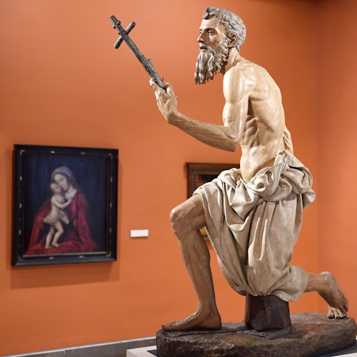Une statue d'un homme barbu tenant une croix se tient à côté d'un tableau encadré d'une femme tenant un enfant dans une galerie.
