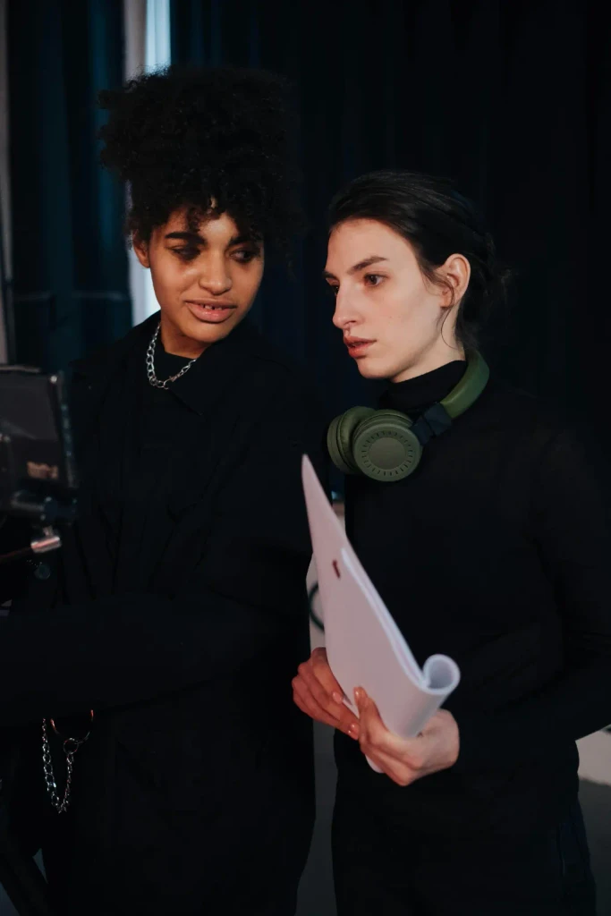 Two women in black focus on a screen in a dark studio. One wears green headphones and holds a white script.