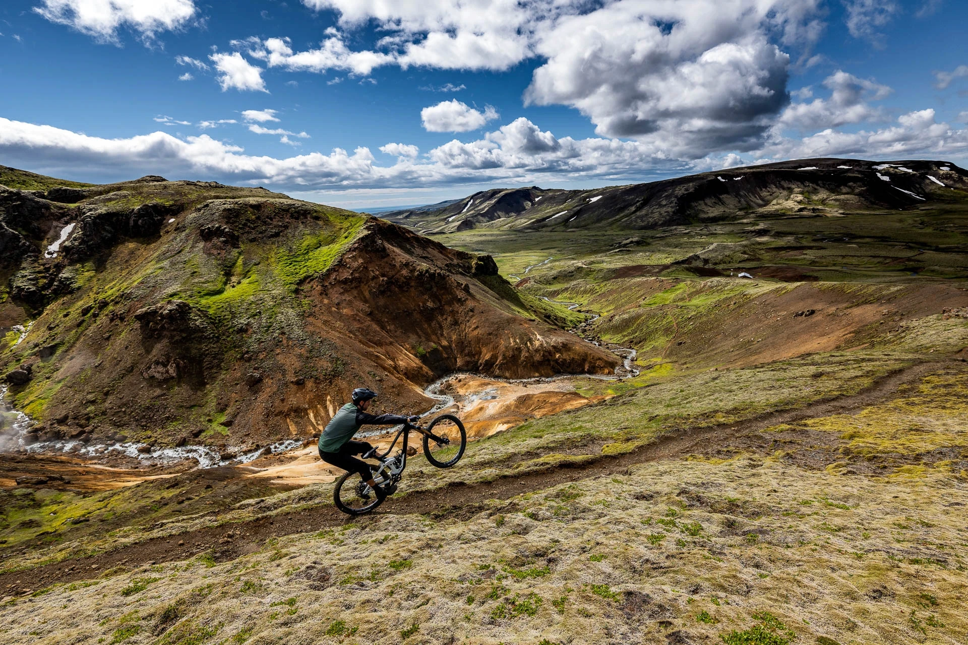 Mountain biker doing a wheelie on an open trail in a wide volcanic valley.