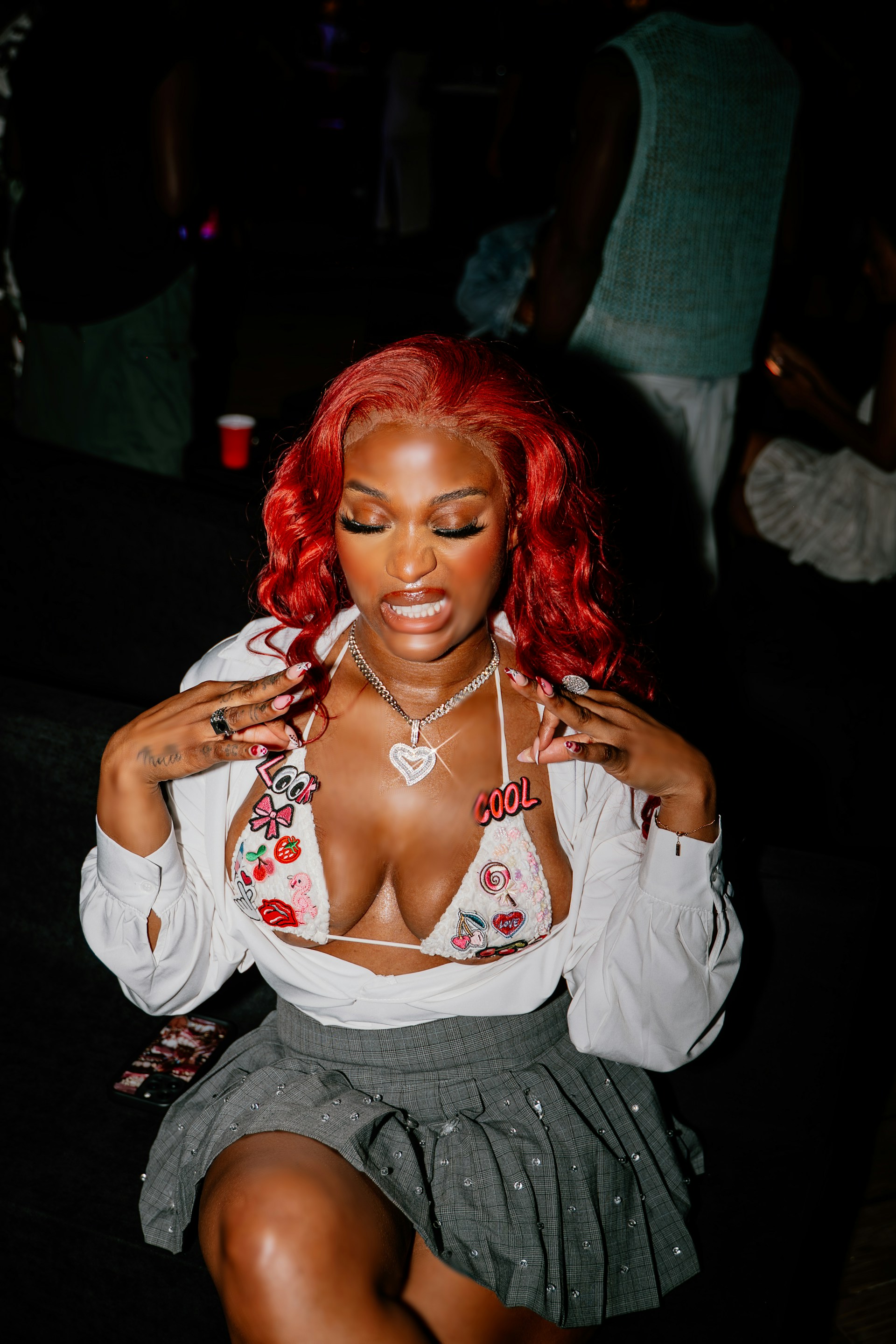 A mid-shot of the woman sitting down, gesturing with her hands near her chest. The white bikini top is adorned with various colorful patches and pins.