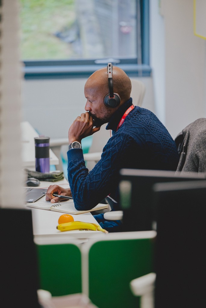 A person sits at a desk in an open office space, wearing headphones and looking at a computer screen. One hand rests on the keyboard while the other is near the person’s face. A jacket is draped over the back of the chair, and a water bottle and pieces of fruit are visible on the desk. A large window in the background lets natural light into the room.