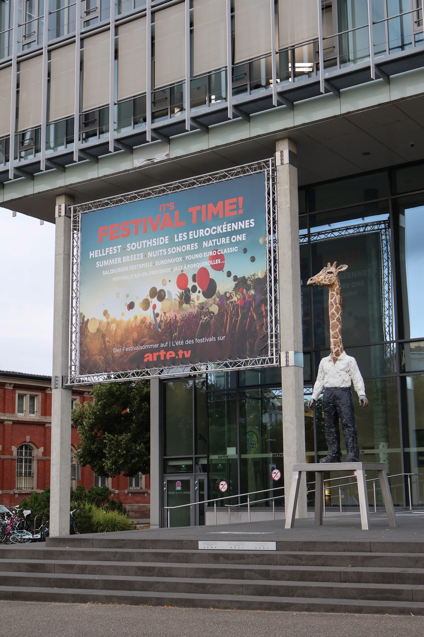 A large banner with the word “Festival Time” and “arte” written on it, in front of a glass façade of a modern building and a giraffe statue standing in front of the building.