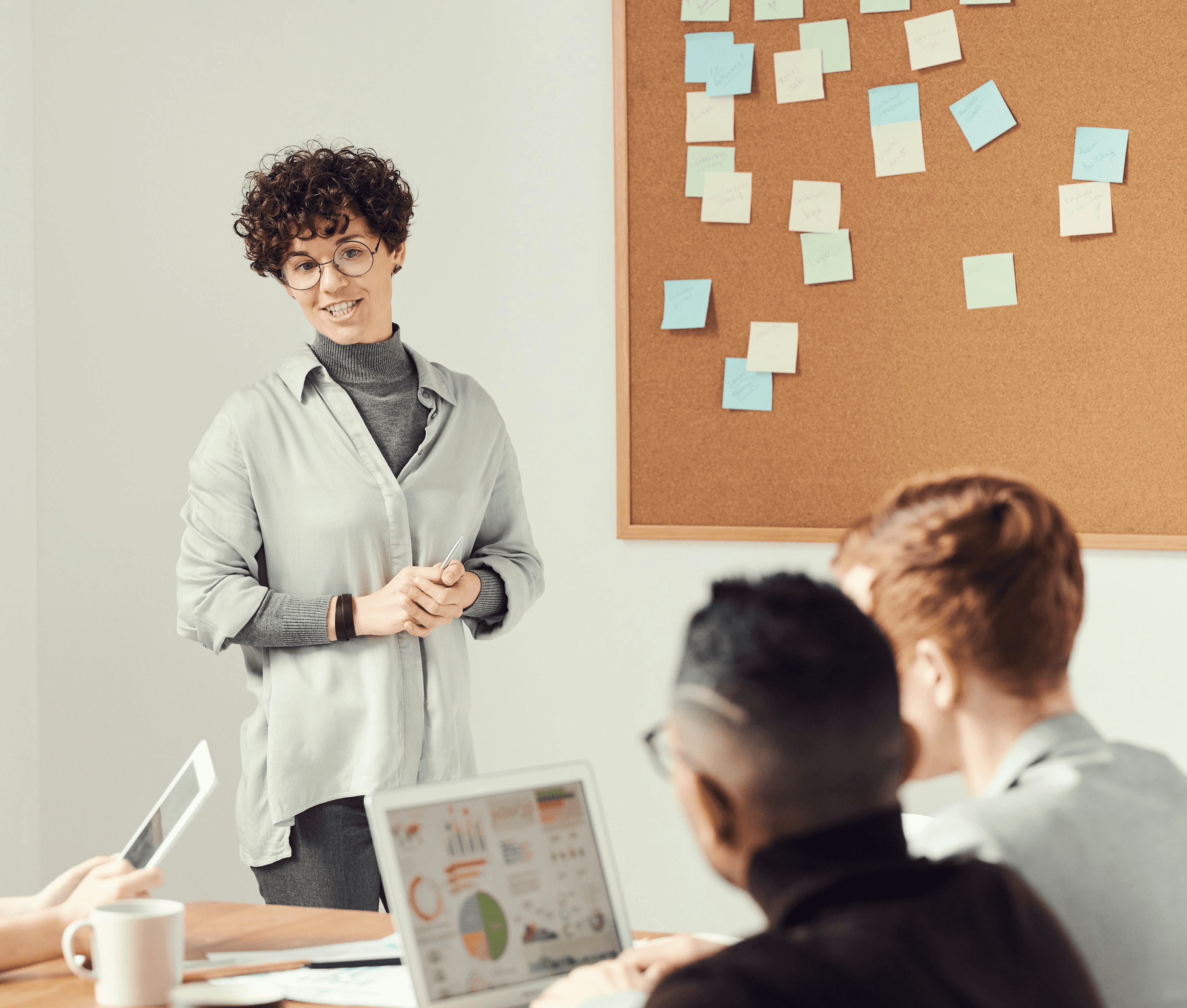 A woman presenting to a small team in a meeting room, with a corkboard and sticky notes in the background.