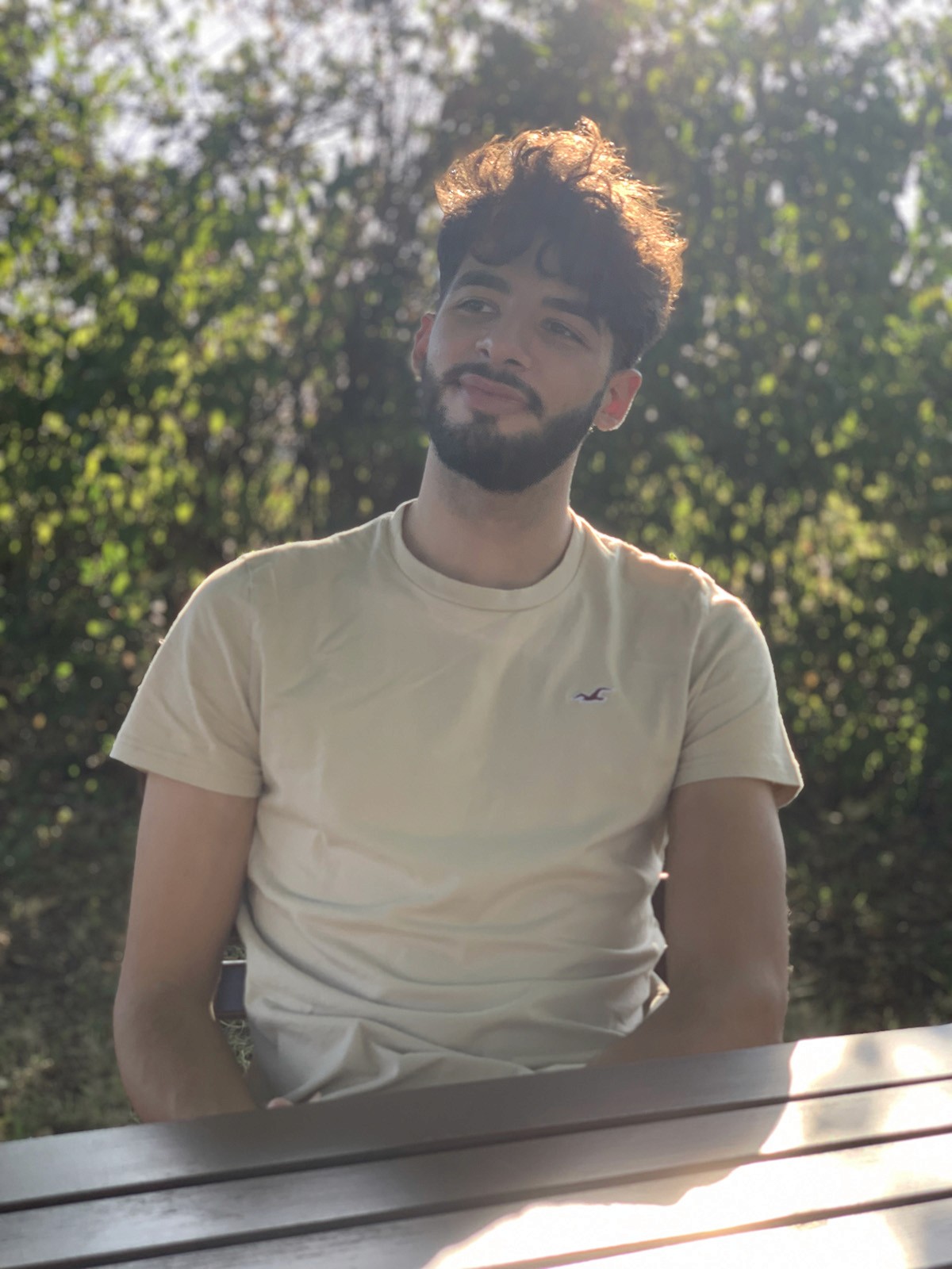 Portraitaufnahme von Saleh Chaaban im kurzen T-Shirt an einem Holztisch in natürlicher Umgebung. Im Hintergrund sind grüne Büsche zu sehen, während Sonnenlicht auf die Person fällt, die entspannt in die Ferne blickt.