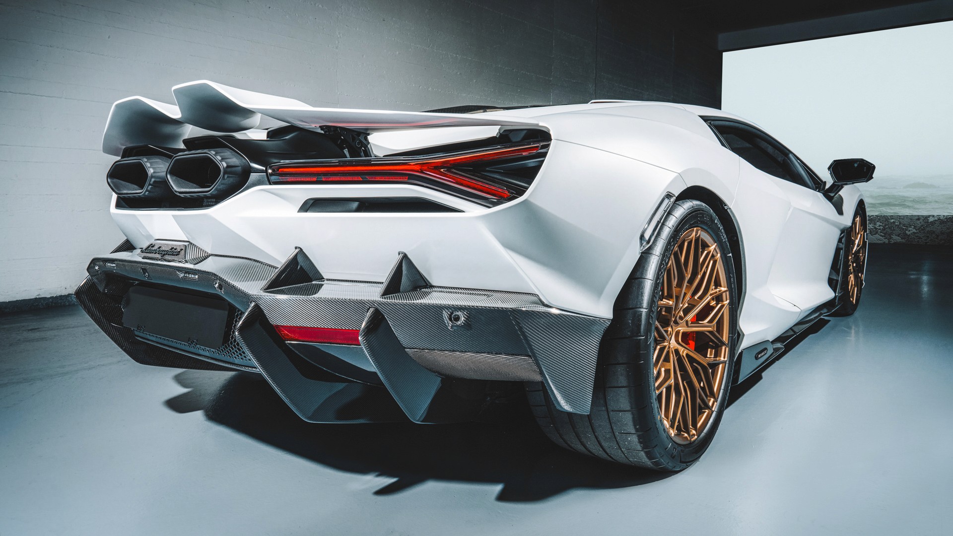White Lamborghini Revuelto with bronze wheels, photographed from the rear three-quarter in a studio, showing the Y-shaped taillights, hexagonal twin exhausts, carbon-fibre diffuser and red brake calipers, on a polished reflective floor.