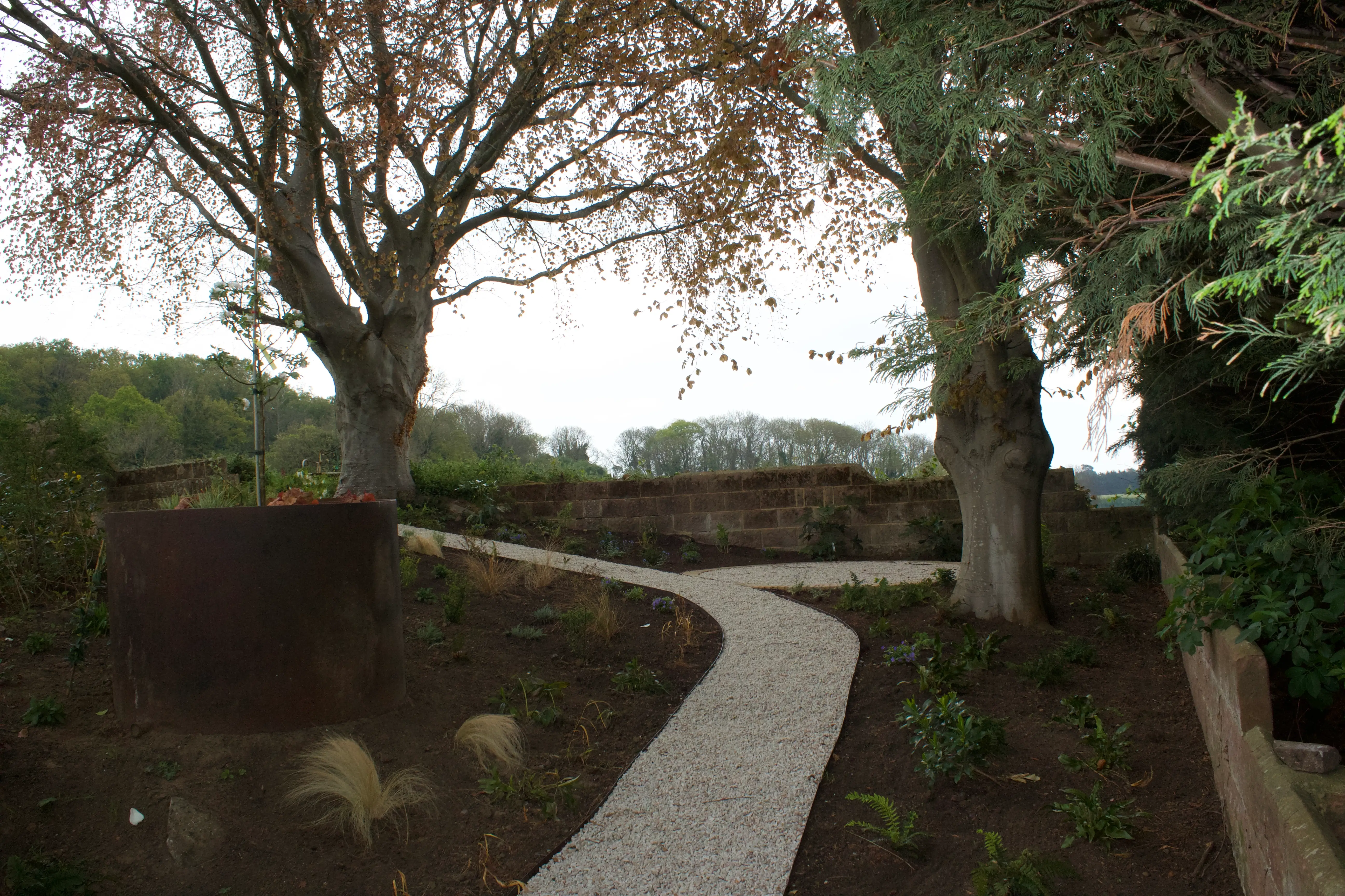 A winding pathway lined with greenery, leading through trees and shrubs towards a cloudy sky.