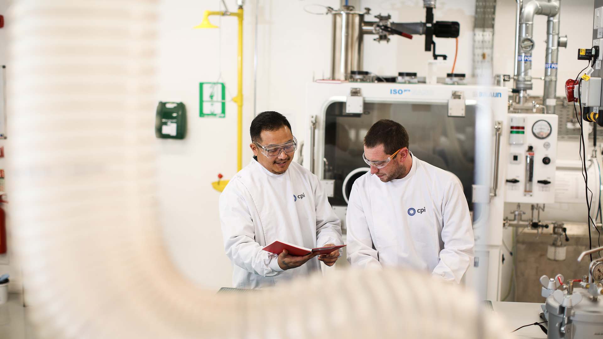 Two men in white CPI lab coats and safety glasses reading a red folder in front of a glovebox and process equipment