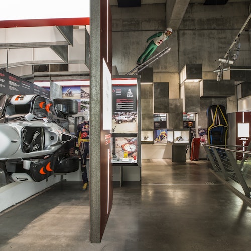 Mostra che presenta un'auto da corsa, cimeli sportivi e un manichino vestito da saltatore con gli sci, in uno spazio museale moderno e industriale.