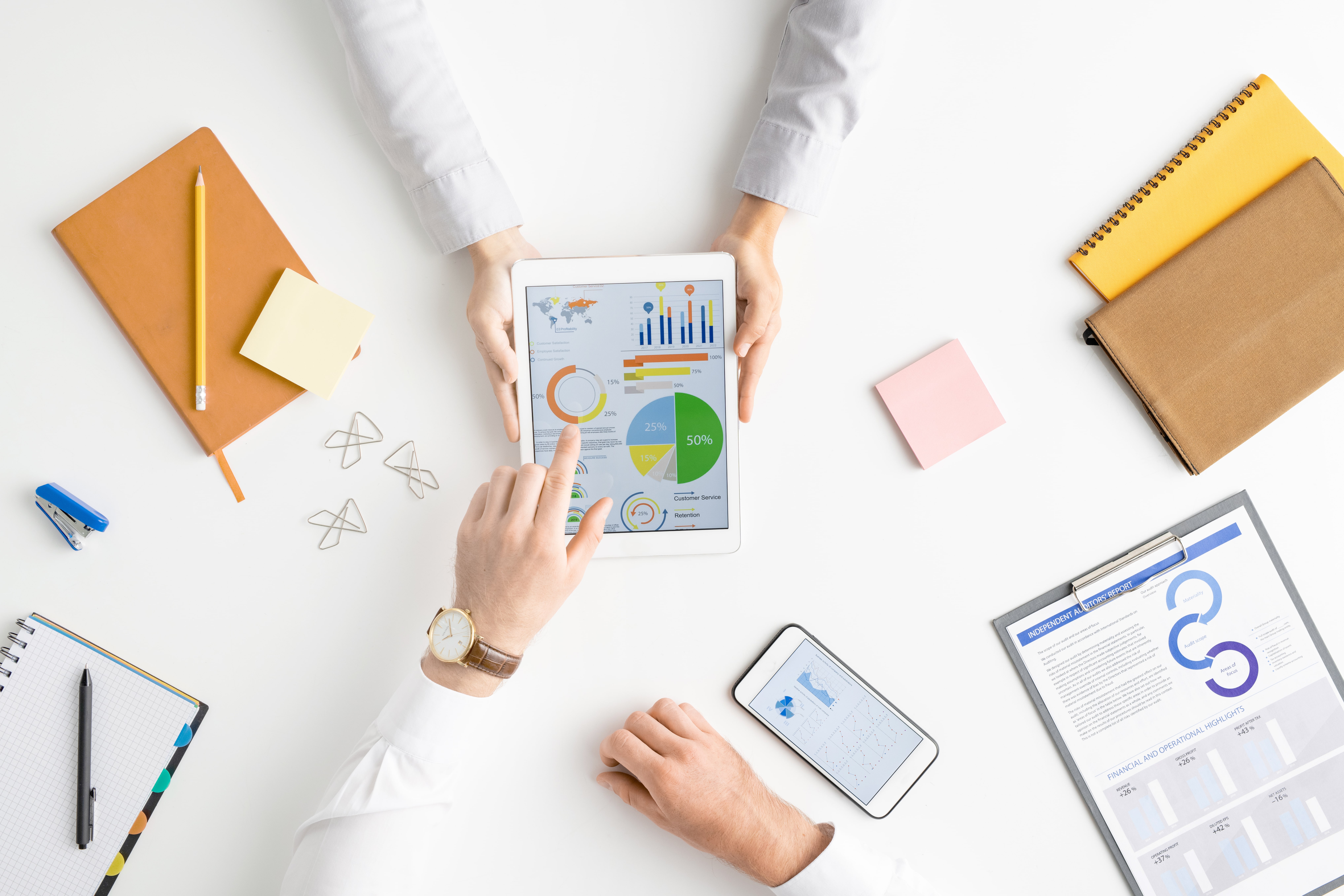 Overhead view of two colleagues at a business meeting, analyzing financial charts and data on a tablet and printed reports on a white office desk.