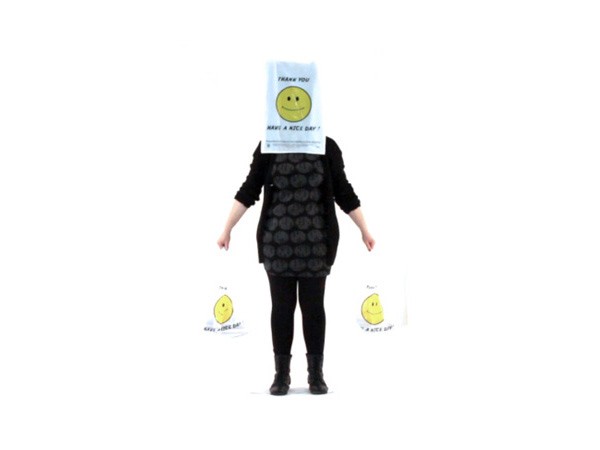 A person stands against a white background, wearing a black top with a pattern of dark circles, black pants, and black boots. Their head is covered by a white bag with a yellow smiley face and the words “THANK YOU” and “HAVE A NICE DAY!” printed on it. In their left hand, they hold two white shopping bags, each displaying a yellow smiley face and the phrase “KEEP CALM AND CARRY ON.”