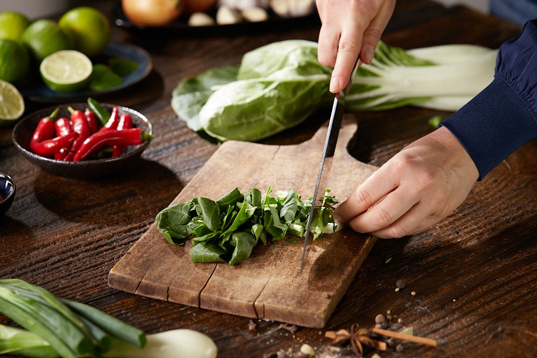 A person is chopping fresh green bok choy on a rustic wooden cutting board, surrounded by various ingredients like red chilies, limes, and green onions on a dark wooden table, capturing a vibrant and organic cooking scene.