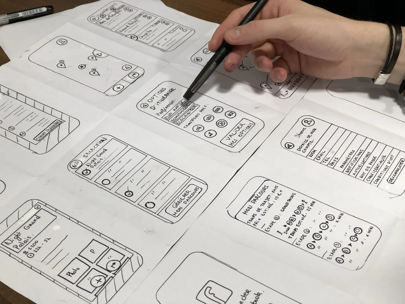 Hand drawn wireframes of a mobile app on several pages spread out on a table with a person holding a pen ready to sketch