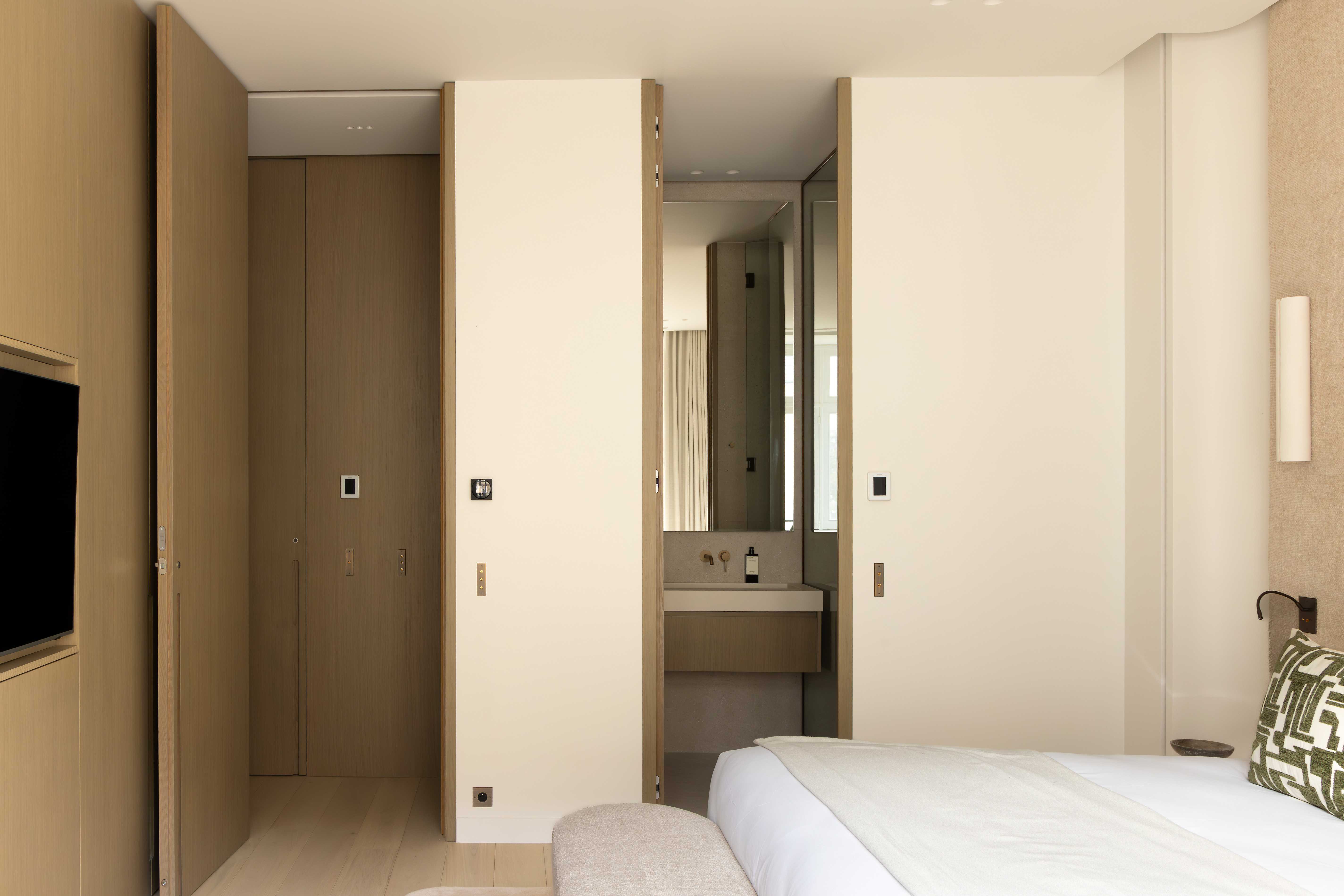 Chambre avec portes ouvertes sur dressing en bois et salle de bain, television murale et tete de lit sable