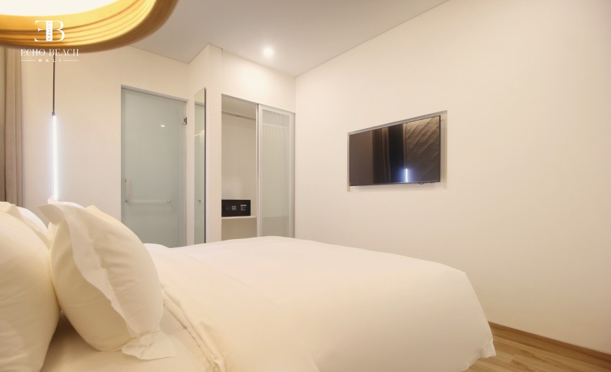 Canggu Two Bedroom Apartment: Second bedroom featuring a comfortable white bed, wall-mounted TV, and sliding door closet with safe inside. Modern amenities at Echo Beach Suite, Bali.