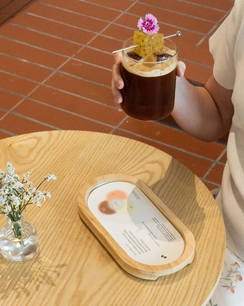 A person holding a glass of coffee topped with a flower and a honeycomb piece, sitting at a wooden table with a small vase of flowers and a menu.