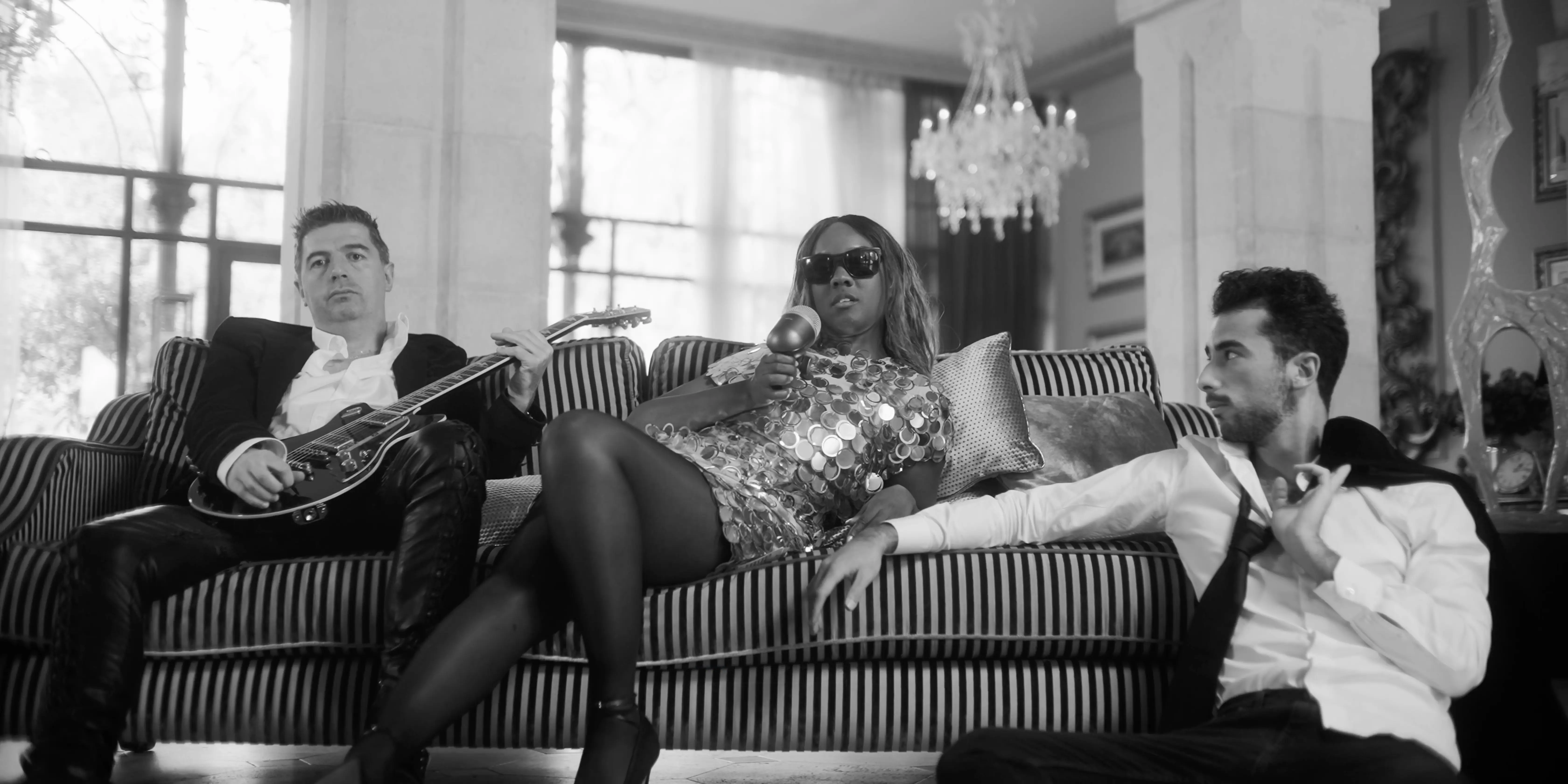 A stylish black-and-white image featuring three elegantly dressed individuals lounging on a striped sofa in a luxurious living room, with one holding a guitar and another wearing sunglasses, exuding a sophisticated and relaxed ambiance.