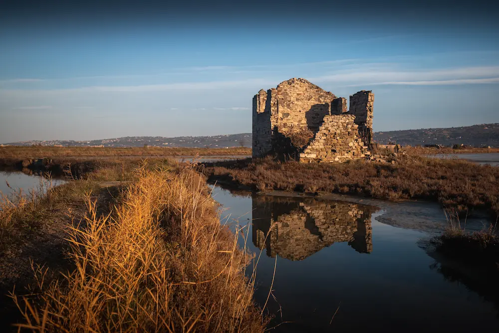 Historic stone ruins at the Sečovlje salt pans in Slovenia, mirrored in calm water with a coastal landscape in the distance.