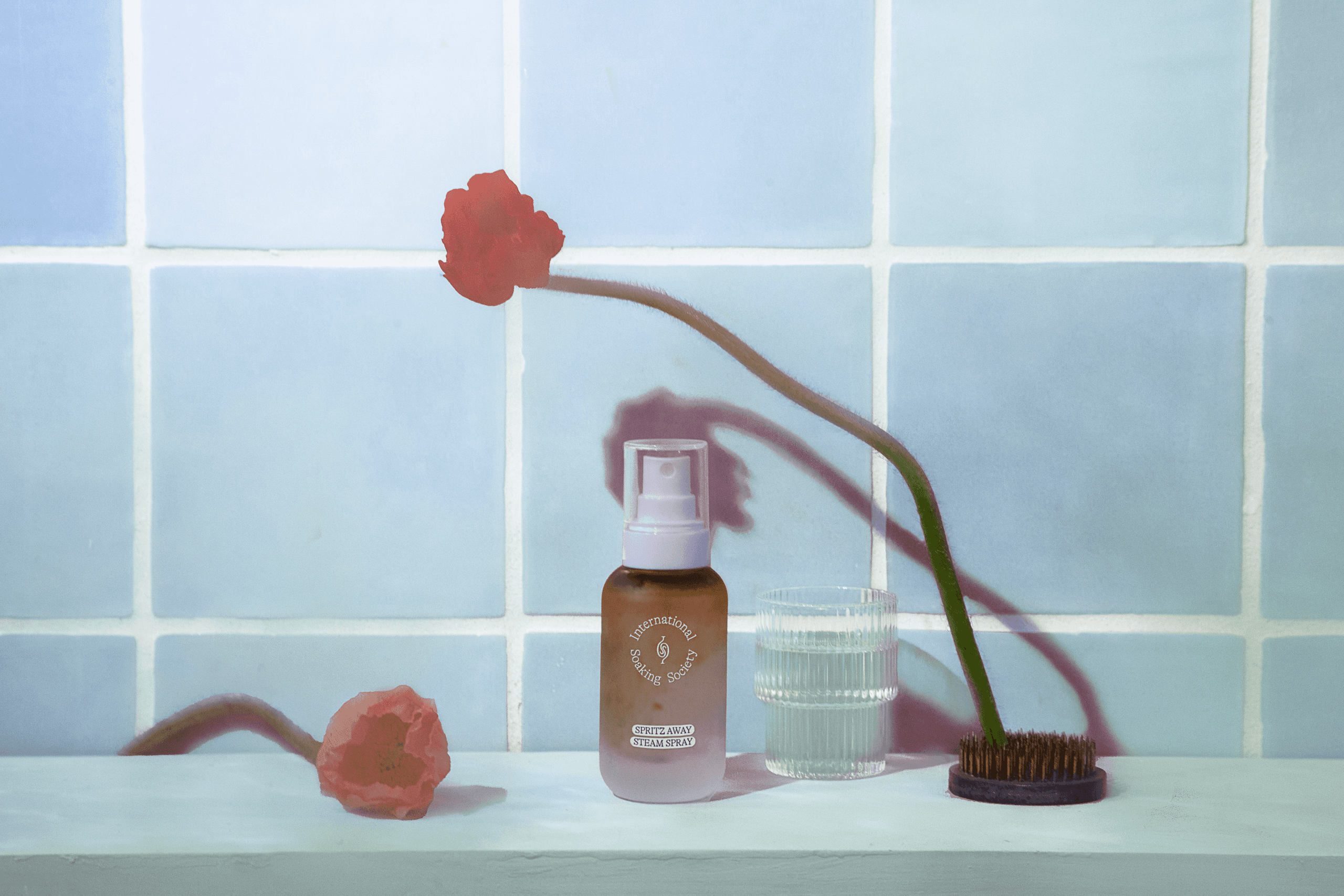 A bottle of Starbucks Starbucks Water Body Mist sits on a white surface against a light blue tiled wall. The bottle is light brown with a white cap and features the Starbucks logo. To the right of the bottle is a clear glass of water and a dark brown hairbrush. A single coral-colored poppy flower lies on the surface to the left of the bottle. The scene is illuminated by natural light, casting soft shadows.
