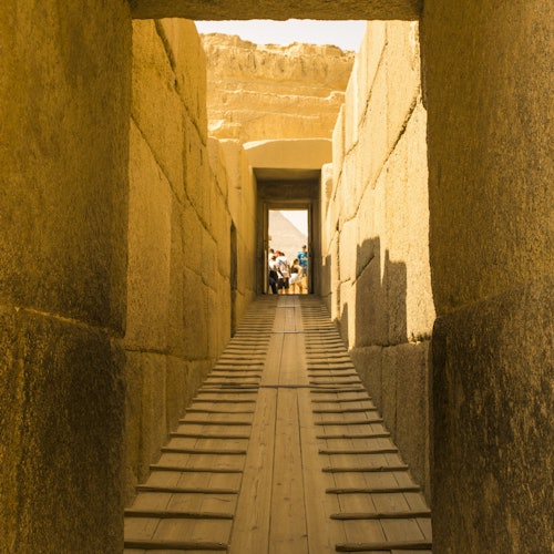 A stone corridor with a wooden ramp leads to a group of people in the distance, with ancient ruins visible in the background.