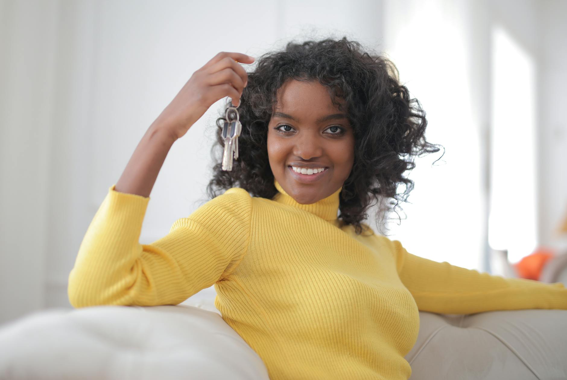 Smiling young woman in a yellow jumper holding up house keys while sitting on a sofa in a bright room