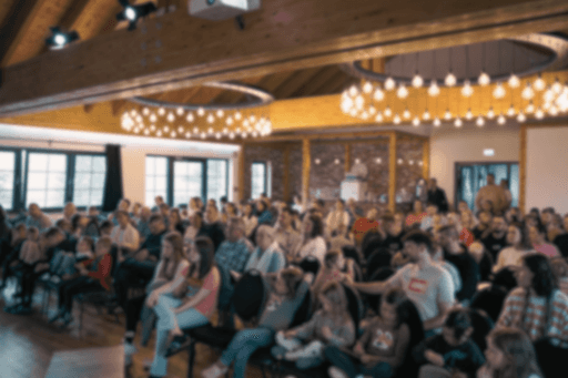 Blick von hinten in einen gut besuchten, gemütlichen Saal mit Holzgebälk und runden Lichterkranz-Leuchtern; viele Menschen sitzen in Stuhlreihen zusammen.