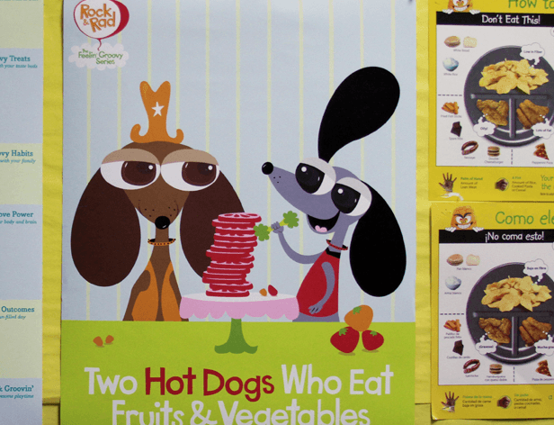 Two cartoon dogs, one wearing a crown and glasses, the other in a red dress, are seated at a table with stacks of red circular objects. The text below reads “Two Hot Dogs Who Eat Fruits & Vegetables.” To the right, a yellow sign displays “Don’t Eat This!” and “Como ele?” with a cartoon dog holding a plate of chips.