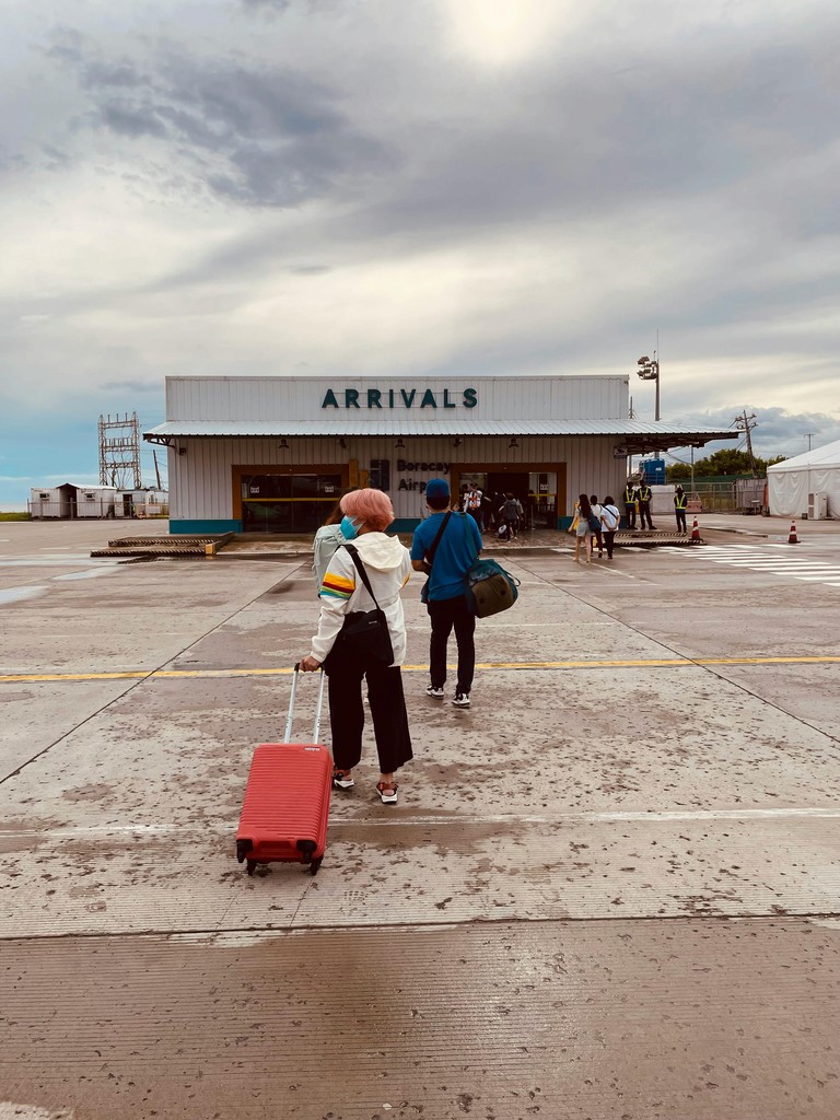Closeup of a group of newcomers to Canada walking outdoors with their suitcases, including a person with pink hair pulling a red suitcase, toward a small "ARRIVALS" building at a Canadian airport terminal, representing the intent to immediately visit the Service Canada desk to obtain their Social Insurance Number (SIN).