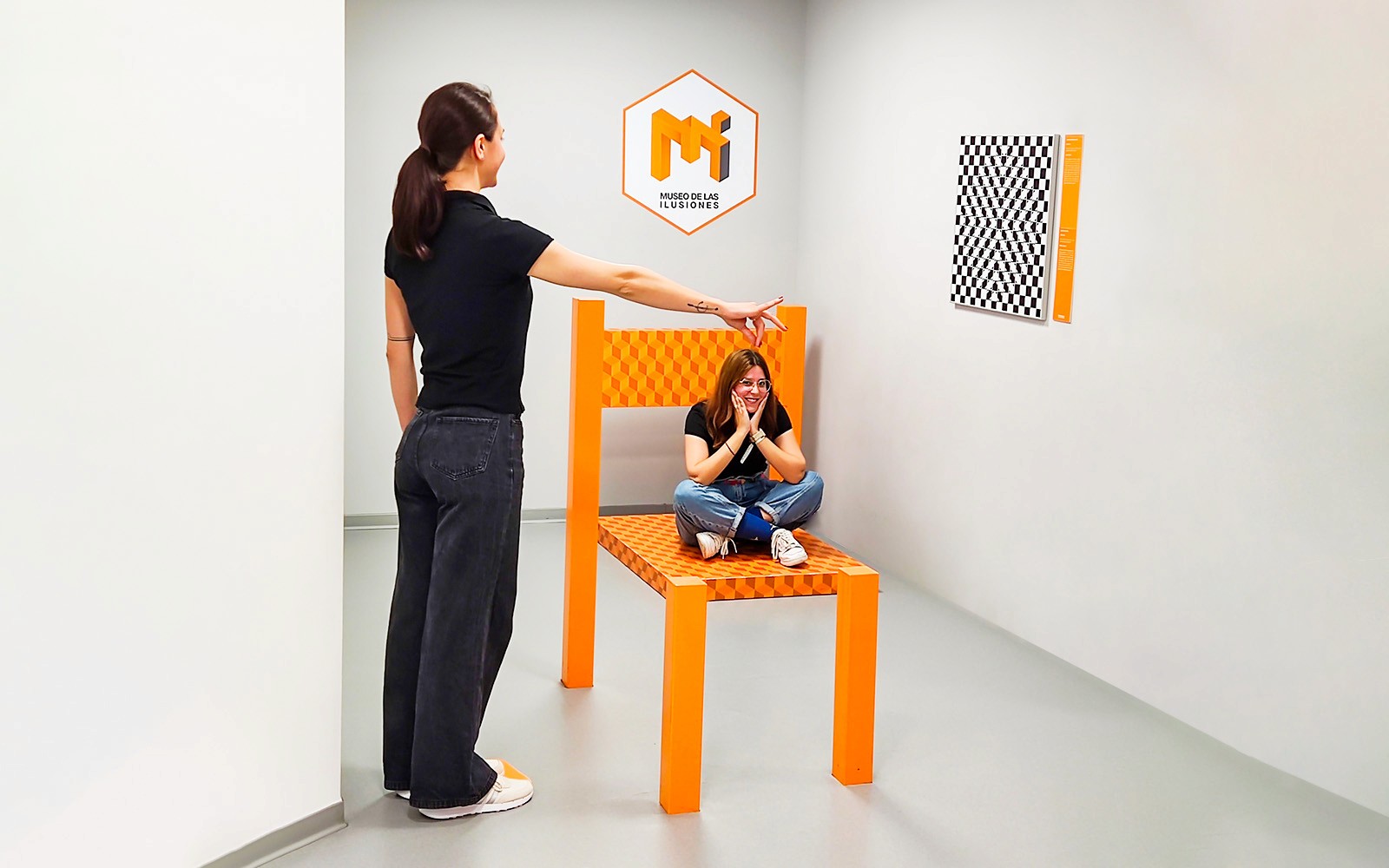 Person pointing at another sitting on oversized chair in perspective room, Museum of Illusions Seville.