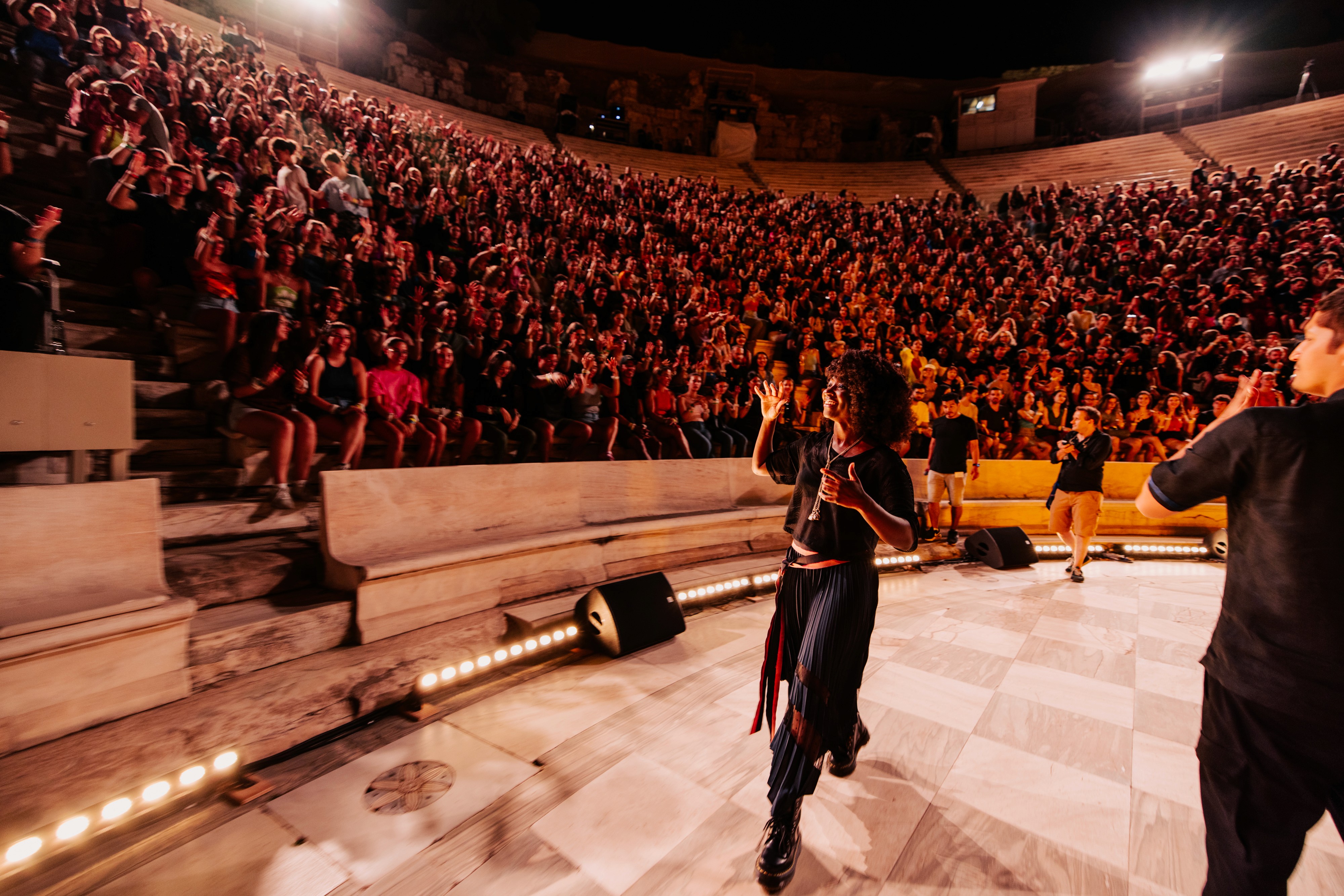 Natasha Ofili (a dark-skinned Black woman) and other performers on stage with a cheering audience in an outdoor amphitheater, The Herodion in Greece, illuminated by red stage lights.