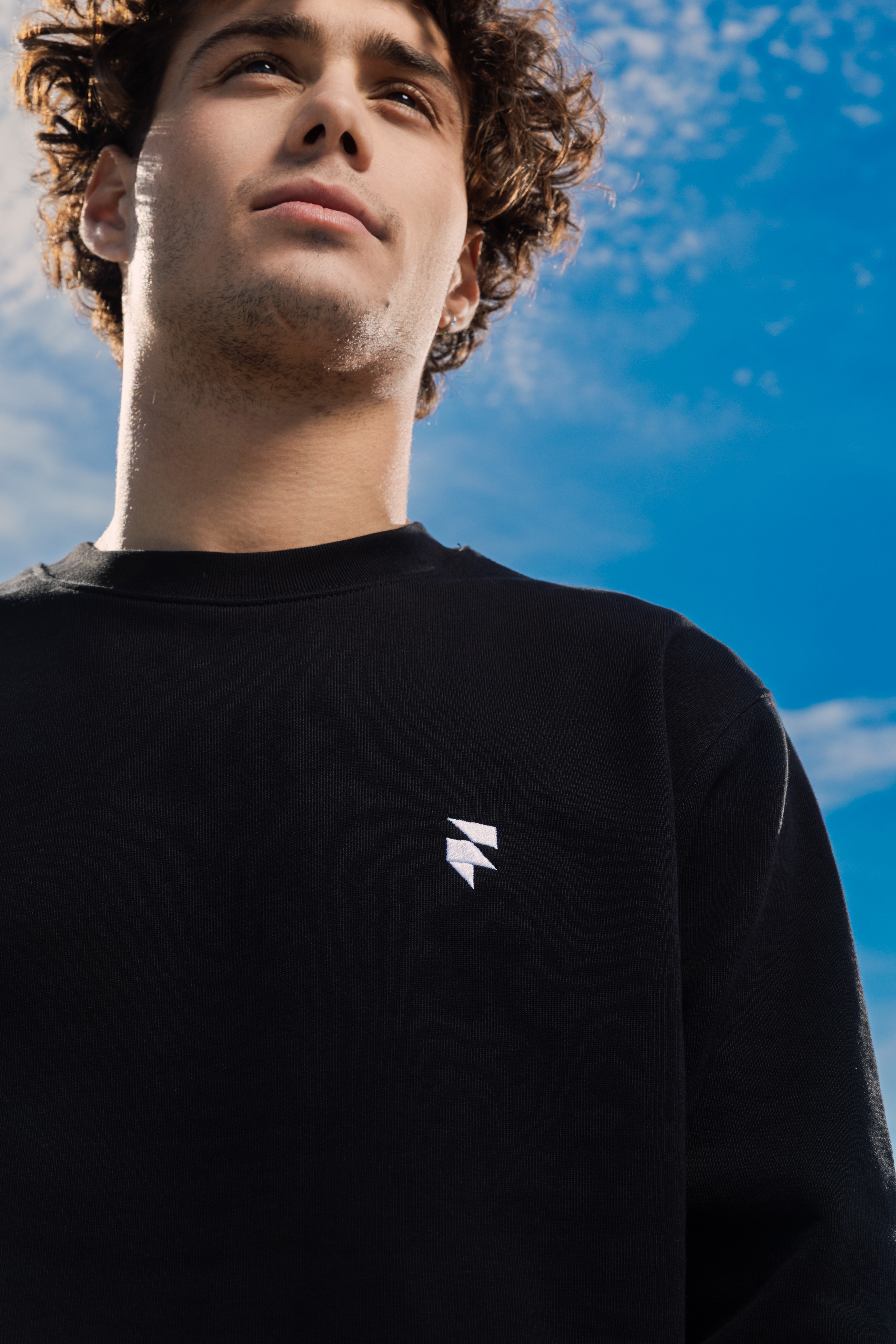 A young man wearing a black sweatshirt with a small white logo stands against a bright blue sky with scattered clouds, looking upwards.