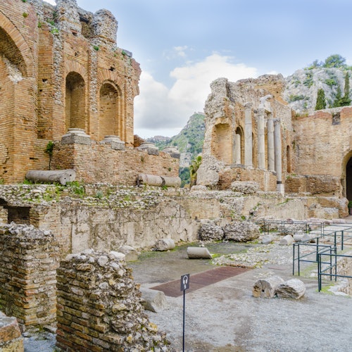 Ruines antiques avec des arches en briques patinées, des colonnes et des débris de pierre épars, le tout sur fond de montagnes.