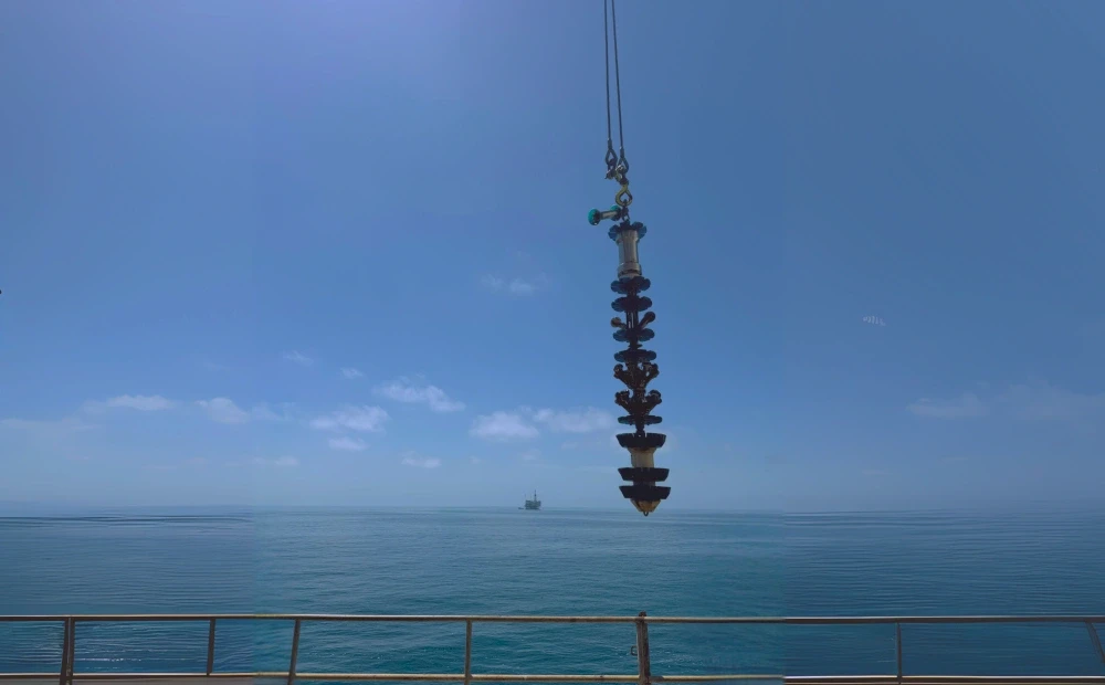 A long, dark cylindrical object suspended from a crane against a clear blue sky and ocean. In the distance, a ship is visible.
