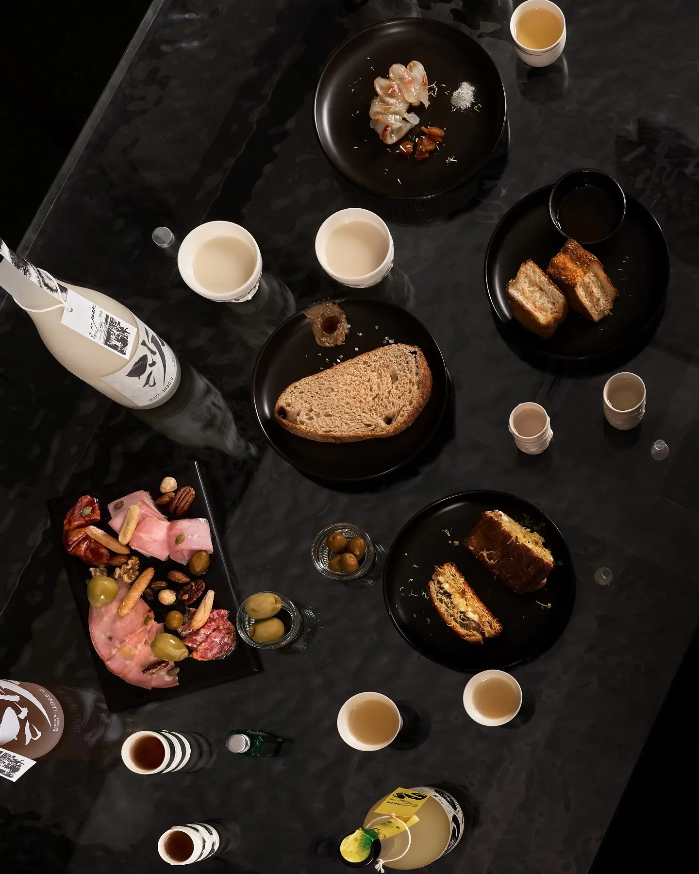 an overhead view of a black table with small sharing plates, bottles of Sahwa traditional liquor, and small glasses filled with makgeolli, fruit wine, and yakju