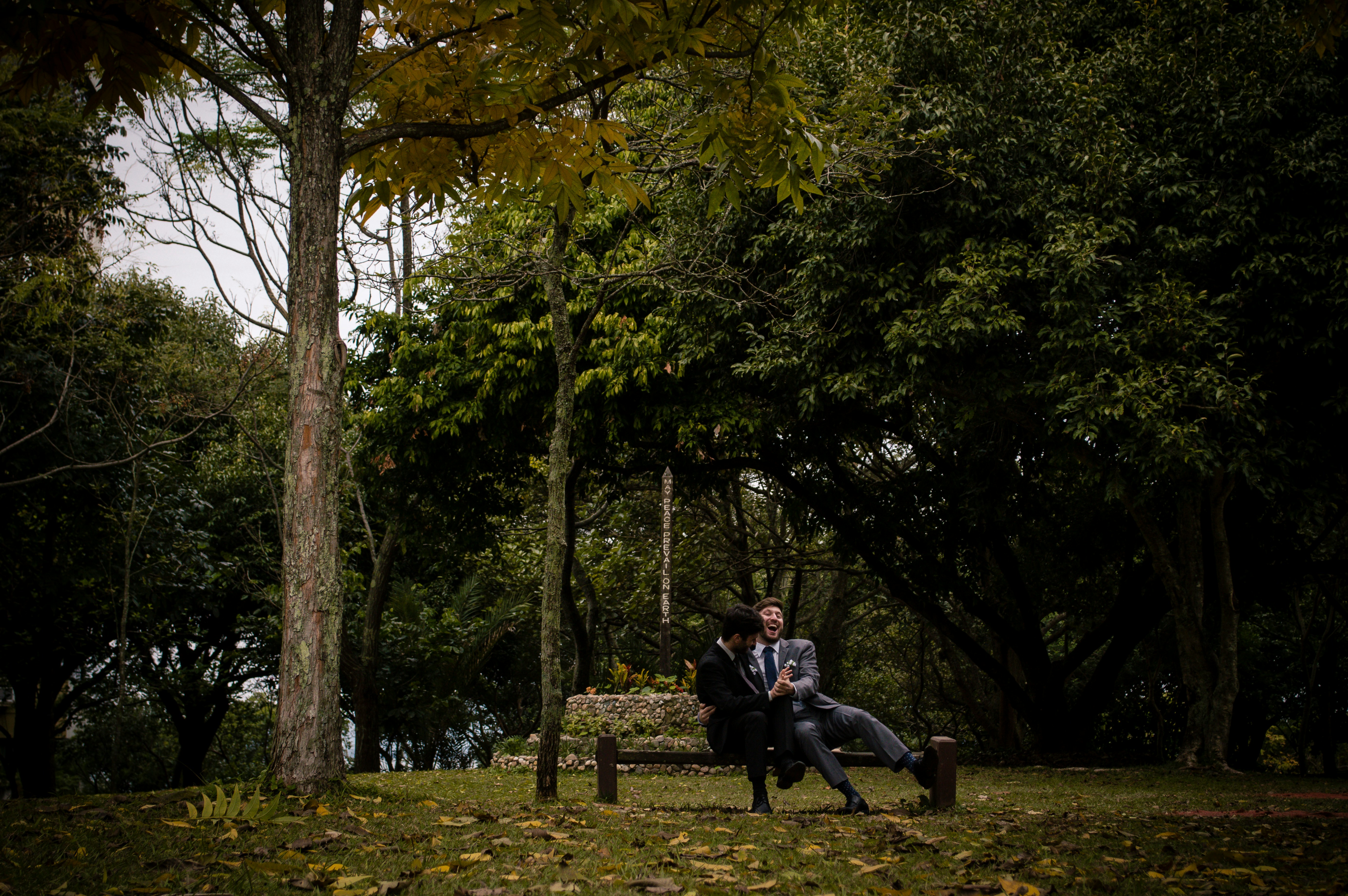 Two men in formal attire share a light-hearted moment on a park bench amidst a lush setting with dense trees and fallen leaves, creating a serene and joyful atmosphere.