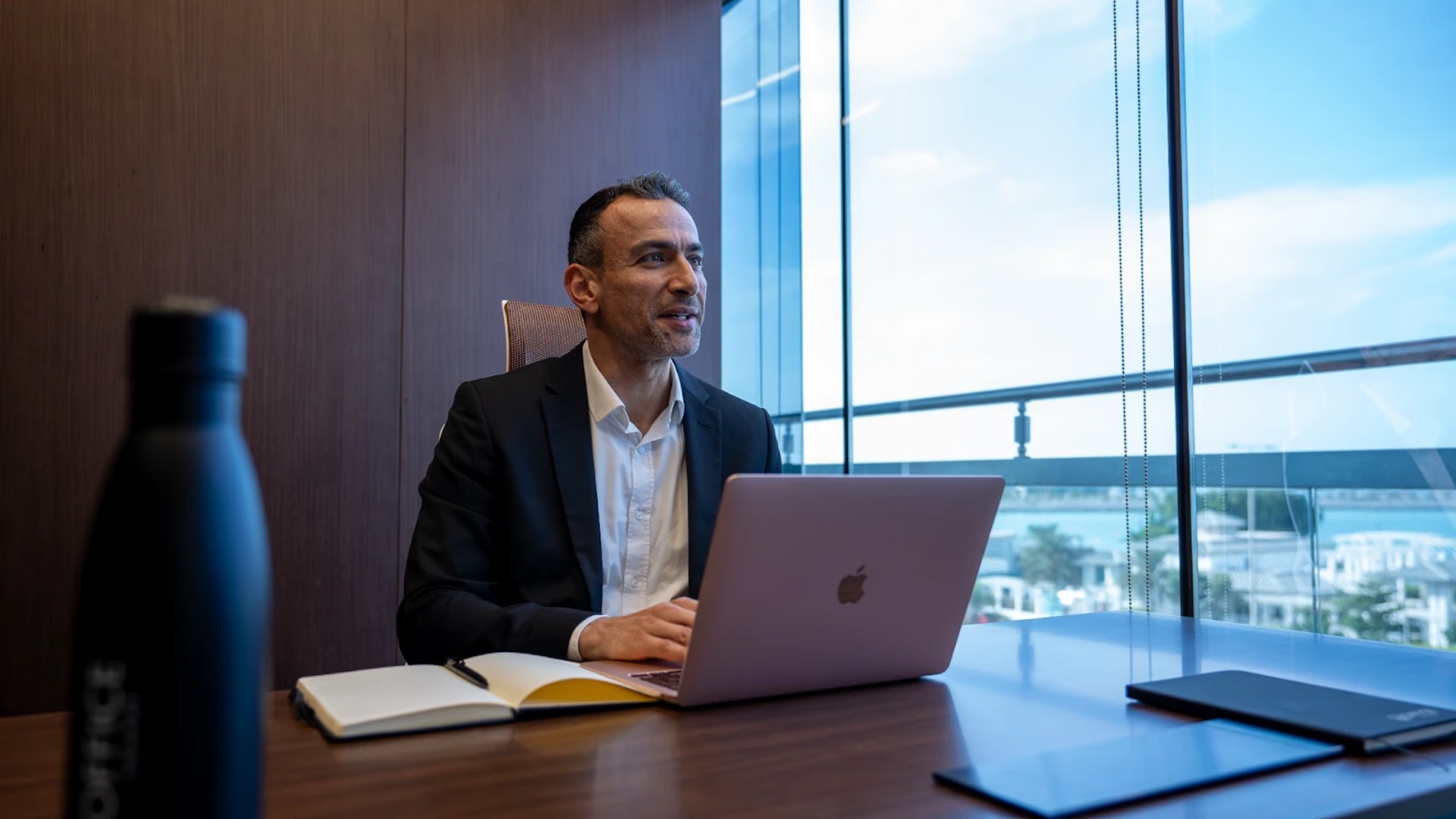   رجل أعمال بملابس رسمية يعمل على مكتبه، مع إطلالة حضارية خلف النافذة توحي بالاحترافية والطموح في بيئة استثمارية مشجعة ومتطورة.
