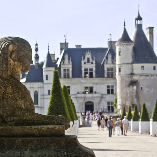 Статуя сфинкса из камня на переднем плане, дорожка, обсаженная коническими кустами, ведущая к большому замку с башенками на заднем плане.