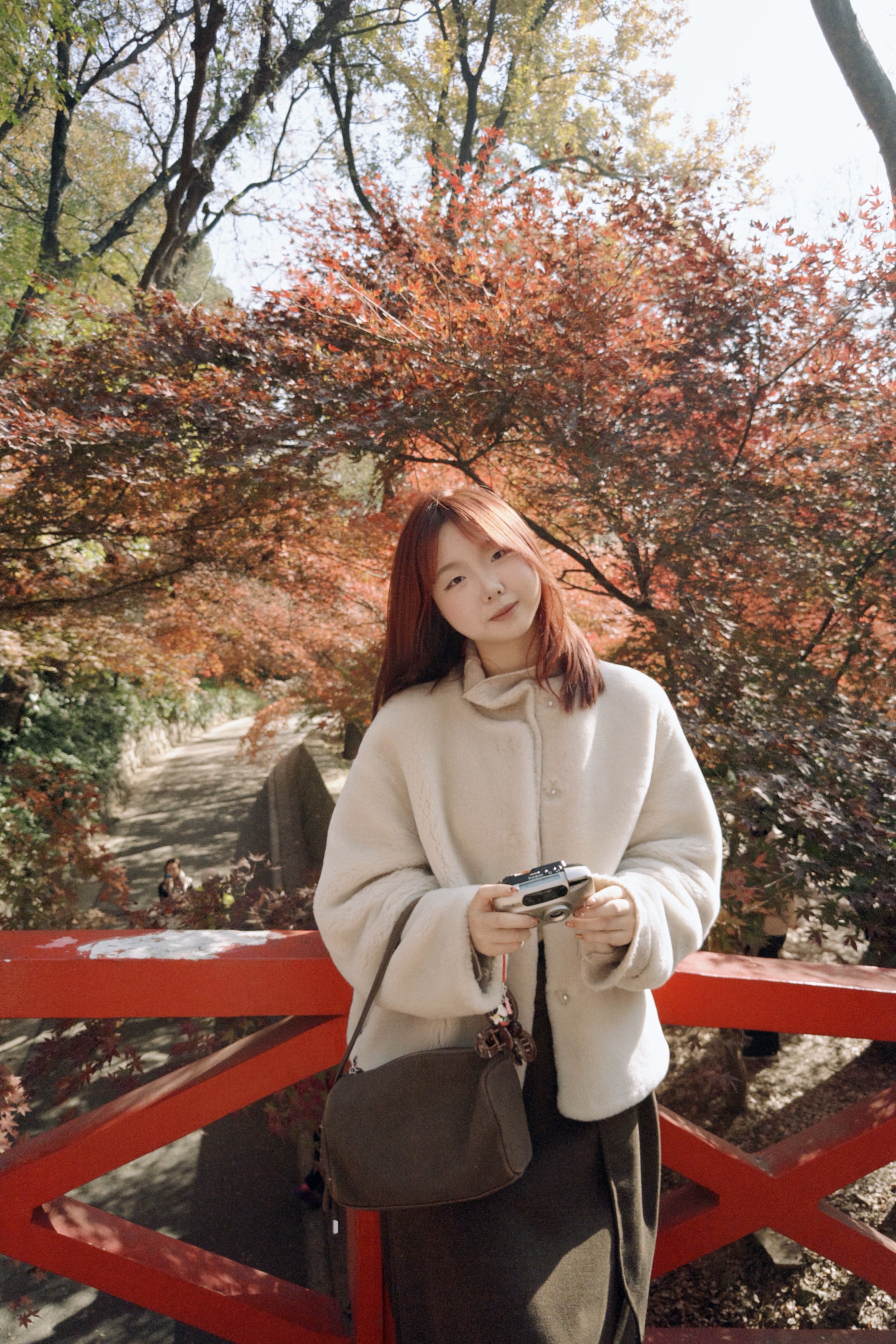 Una persona con una camiseta negra y pantalones de color claro está de pie en la cima rocosa de una montaña al atardecer, sosteniendo una cámara frente a su cara para tomar una foto. Un mar de nubes se extiende detrás de ellos, iluminado por la luz dorada del sol poniente.