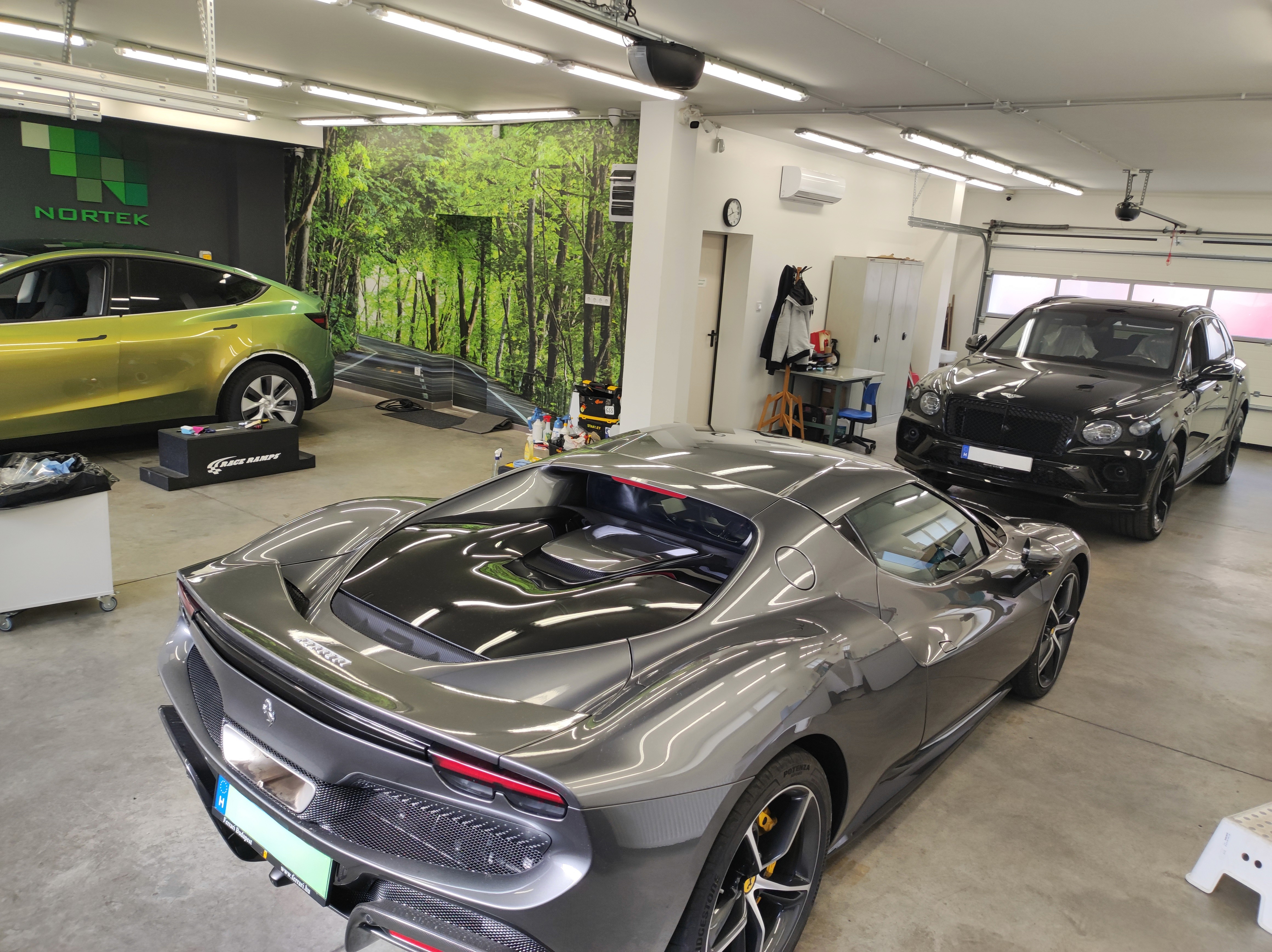 Szürke Ferrari 296 GTB karosszériavédő fóliázása a háttérben egy fekete Bentley Bentayga PPF fóliázásra felkészítve a Nortek műhelyében.