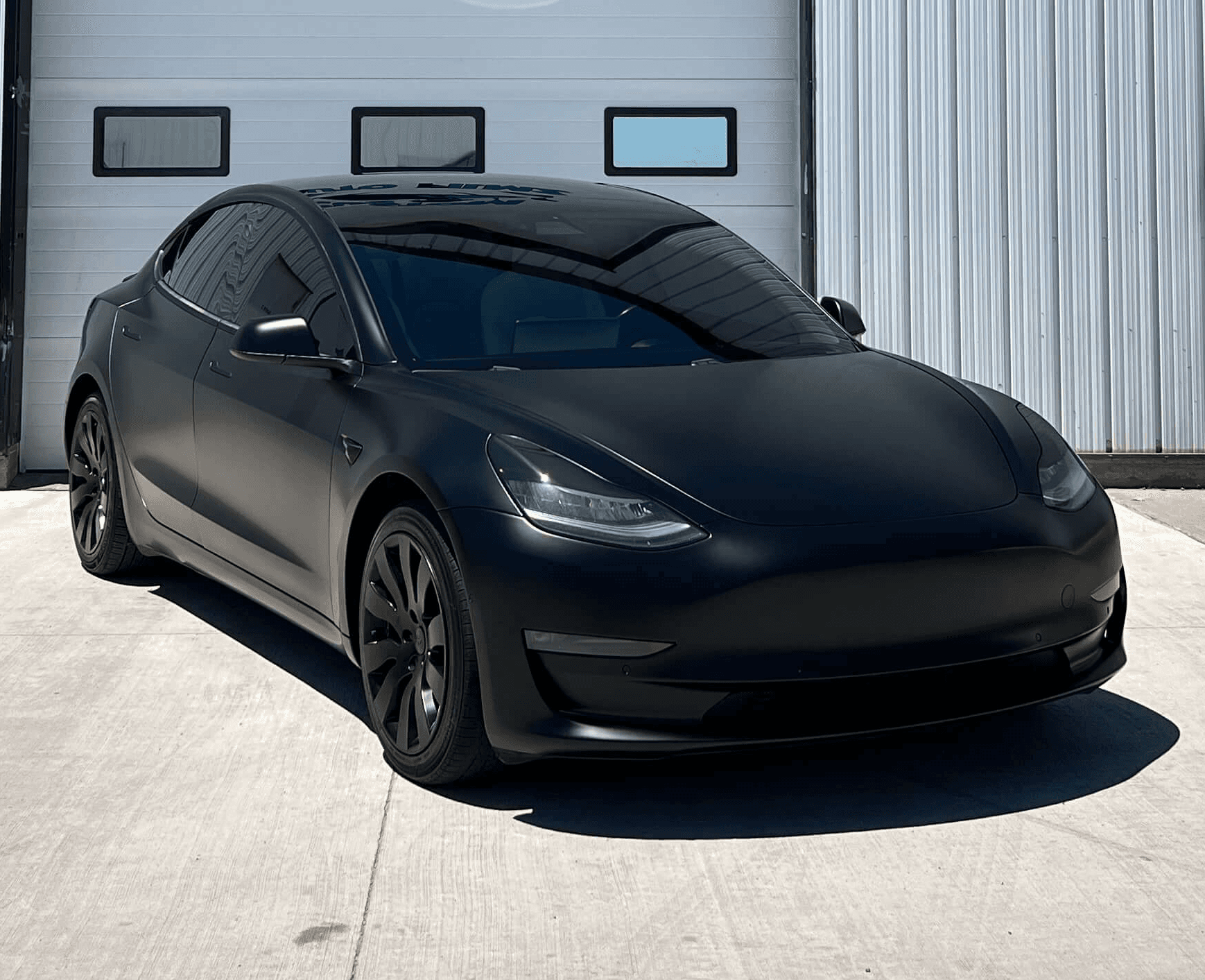 A matte black Tesla Model 3 is parked on a concrete driveway in front of a white industrial building.