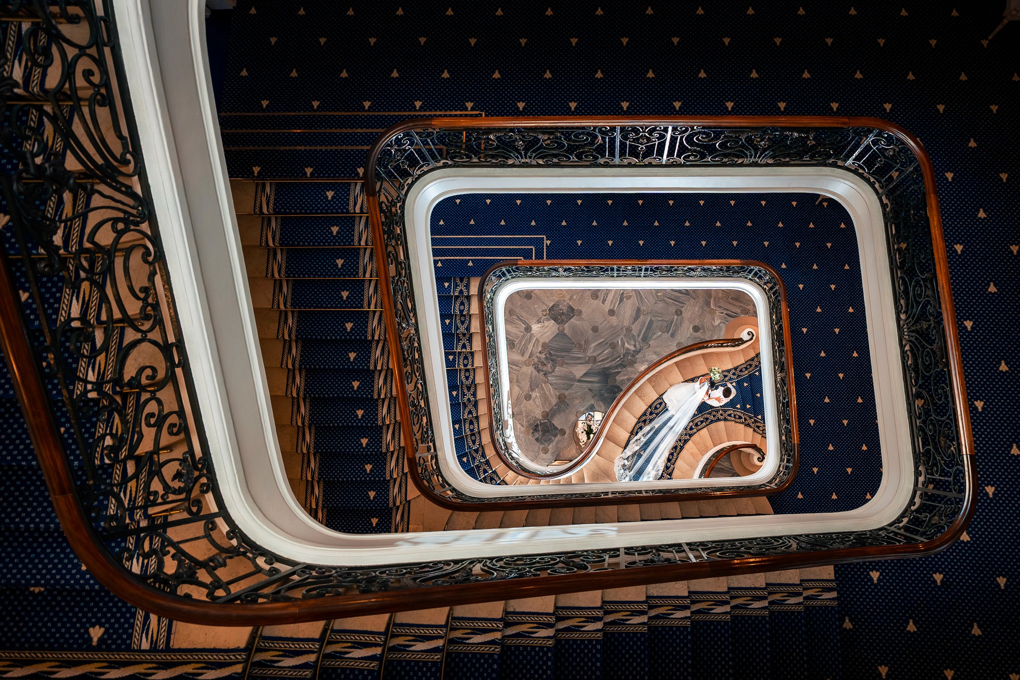 Bride and groom descending the iconic staircase of the Hôtel du Palais in Biarritz, seen from above, framed by elegant curves, symmetry, and refined architectural details.