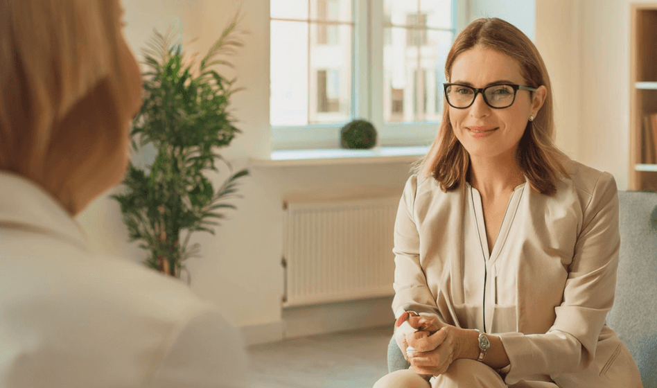 Woman with glasses sitting in a chair, hands folded, engaged in a conversation with another person.