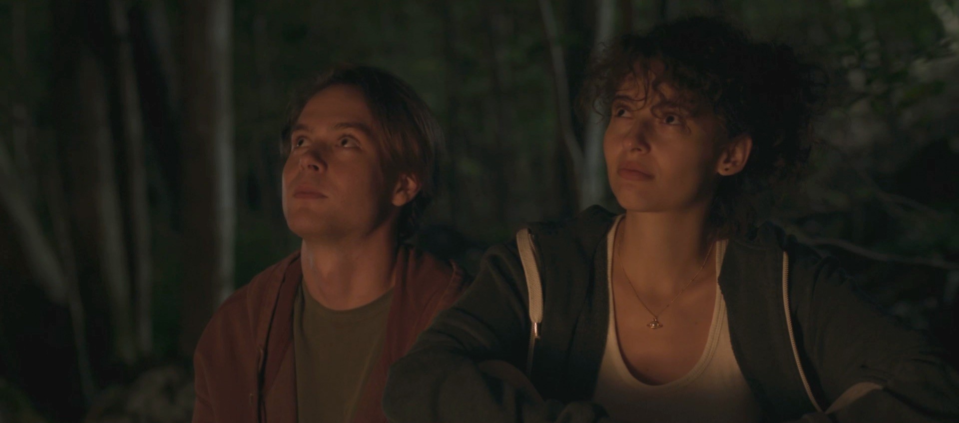 Still from The Floaters: A young man and woman sit side by side, gazing upwards thoughtfully in a dimly lit, forested setting.