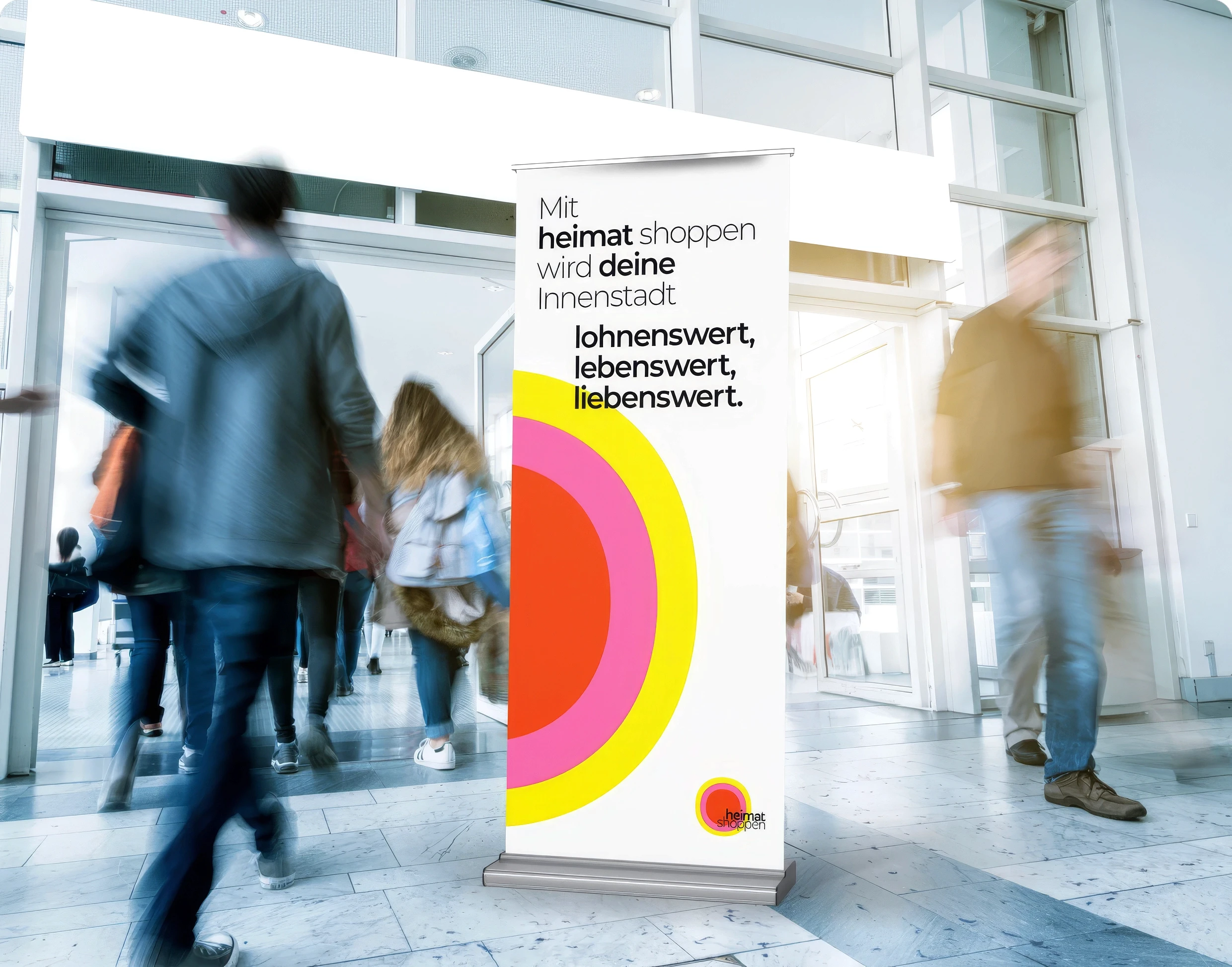 Roll-up-Banner der Kampagne ‚heimat shoppen‘ in einer Eingangshalle mit vorbeilaufenden Passanten und Slogan zur Unterstützung der Innenstadt.