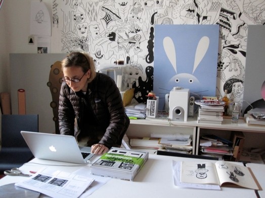 A photo of a person working at a laptop and around her there are sketchbooks, a speaker system and some drawings on the wall.
