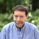 A blue shirted man portrait with a moustache in front of a green plants background