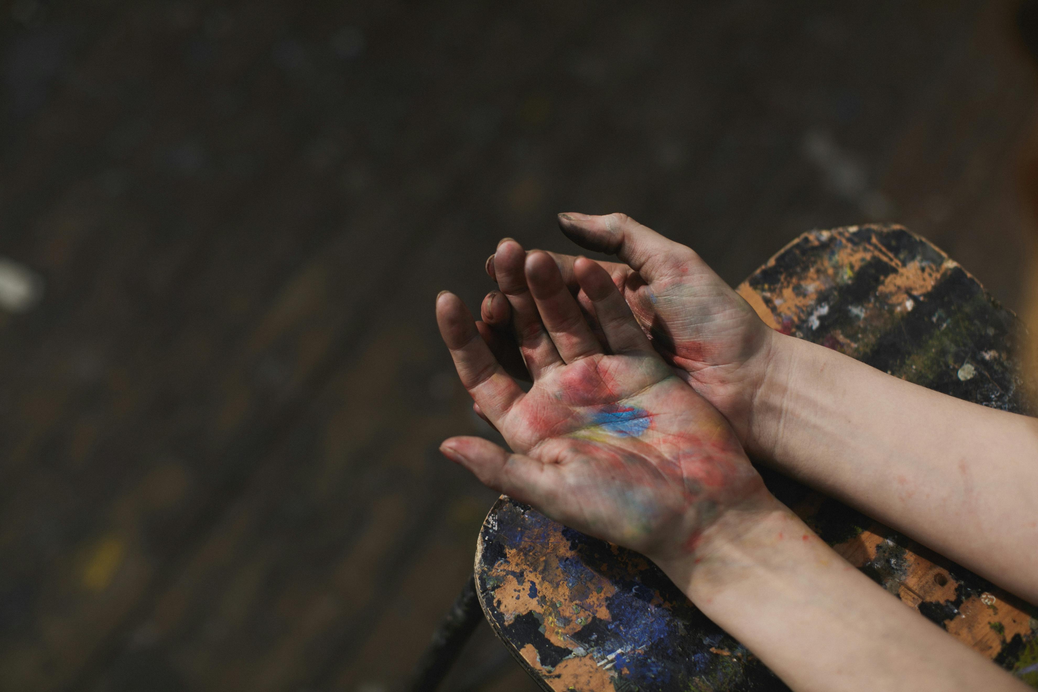 Hands covered with paint, laying across a paint-covered palette with a dark background