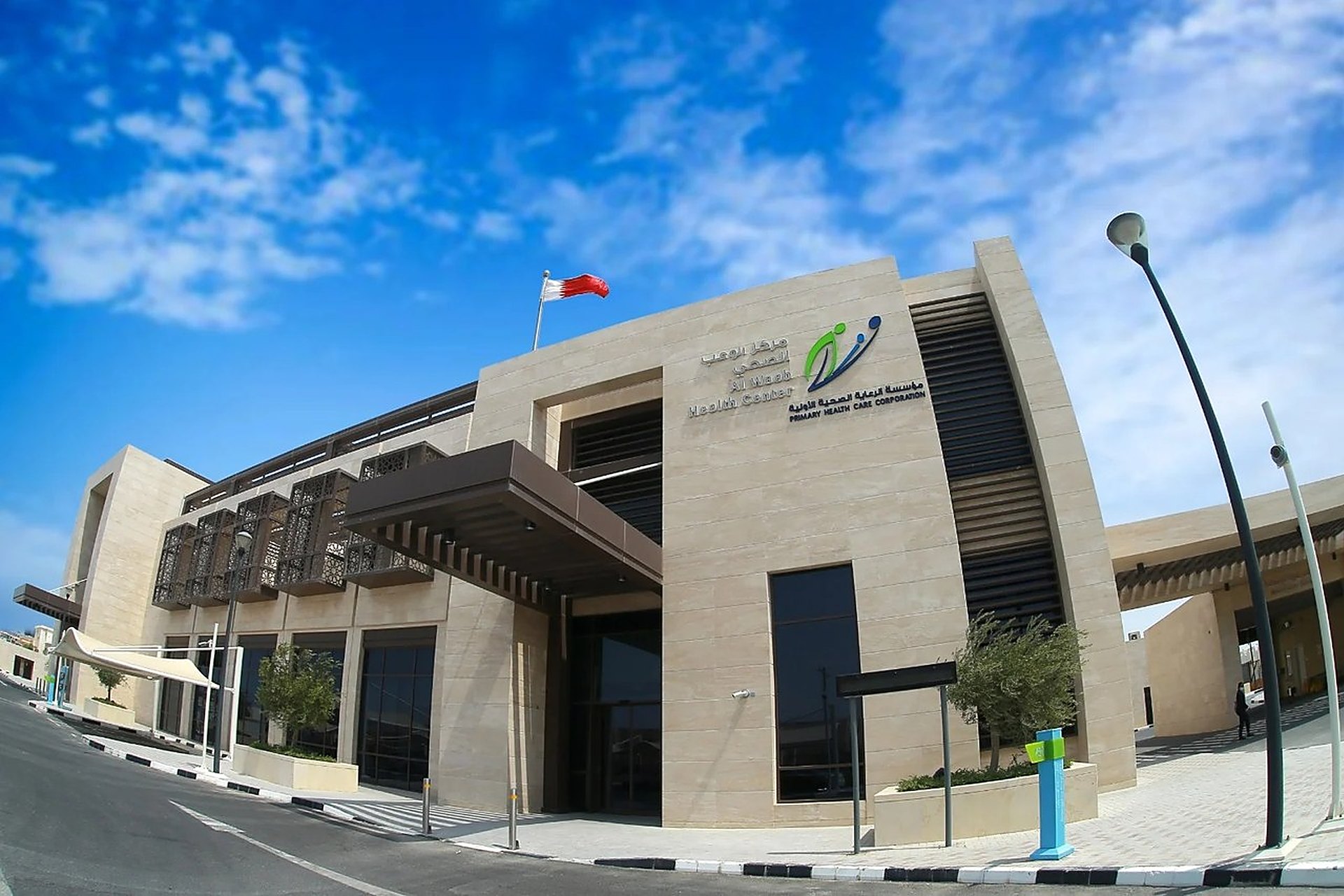 La façade en pierre claire du centre de santé d'Al Waab à Doha, avec son enseigne et le drapeau du Qatar sous un ciel bleu